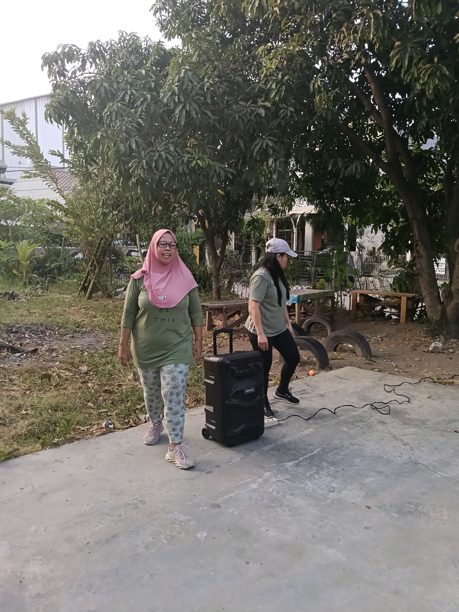 In Jatimulya, Indonesia, two women are outdoors on a concrete surface. The woman on the left, wearing a pink hijab and glasses, is dressed in an olive green long-sleeved shirt with the text "DO THIS" printed on it and patterned leggings. She is smiling and walking towards the camera. To her right, a second woman, wearing a grey baseball cap, is dressed in an olive green t-shirt and black leggings. She is walking away from the camera, looking to her right. A large black portable speaker with wheels is positioned between the two women. Wires are seen on the ground near the speaker. The background features trees, vegetation, and a building with a metal roof. The ground is a mix of concrete and dirt with some dry leaves.