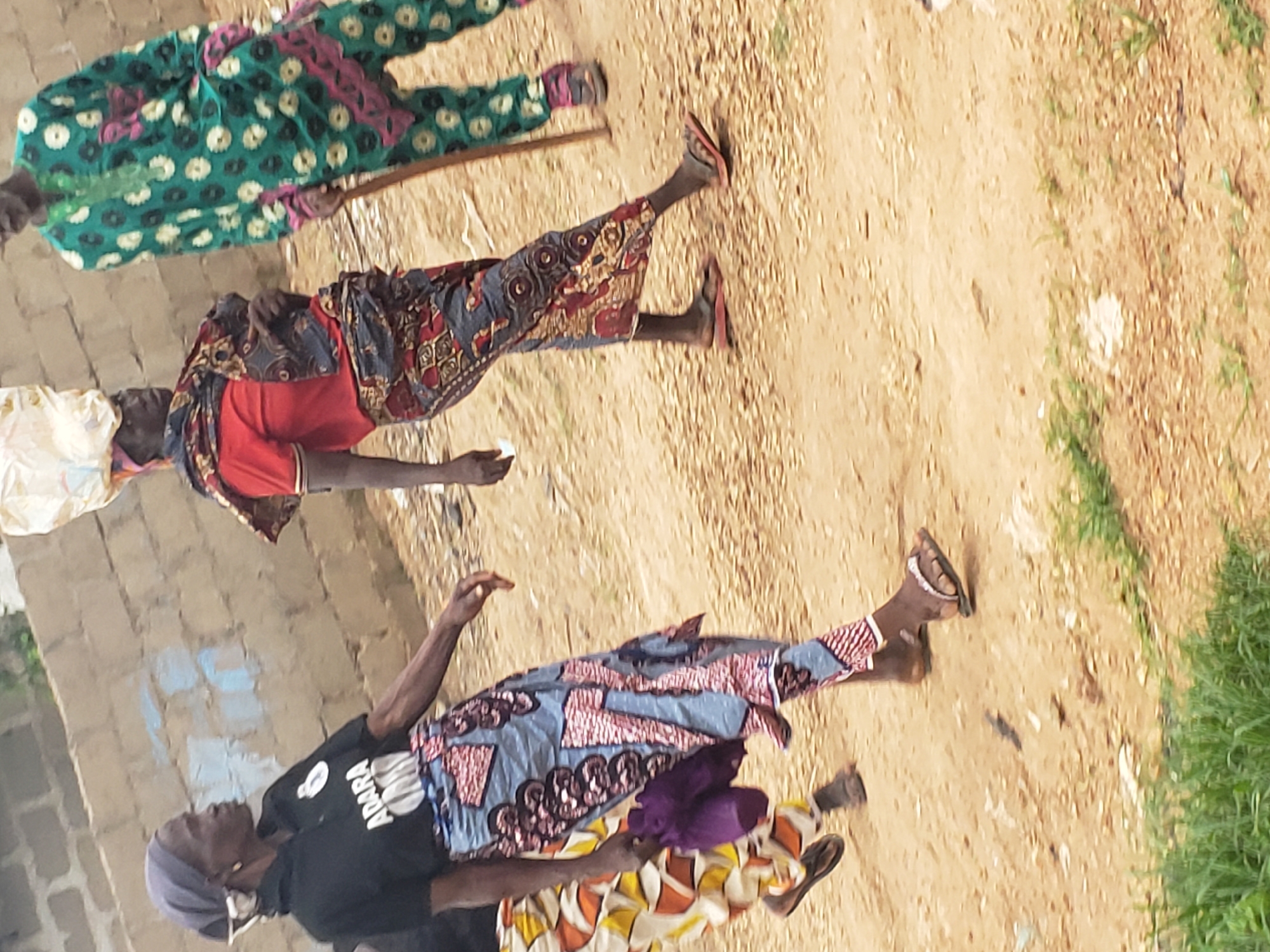 In Kachia, Nigeria, on a dirt road, three individuals walk. The person on the left, wearing a black t-shirt with "ADARA" printed on it, a blue and pink patterned skirt, and a purple headscarf, carries a purple bag. To their right, another individual in a red shirt, a patterned wrap skirt, and a white bundle on their head, walks with a stick. On the far right, a third person sports a green outfit adorned with floral patterns and a pink trim, also holding a stick. The ground is sandy and strewn with debris, bordered by sparse green grass and a brick wall in the background.