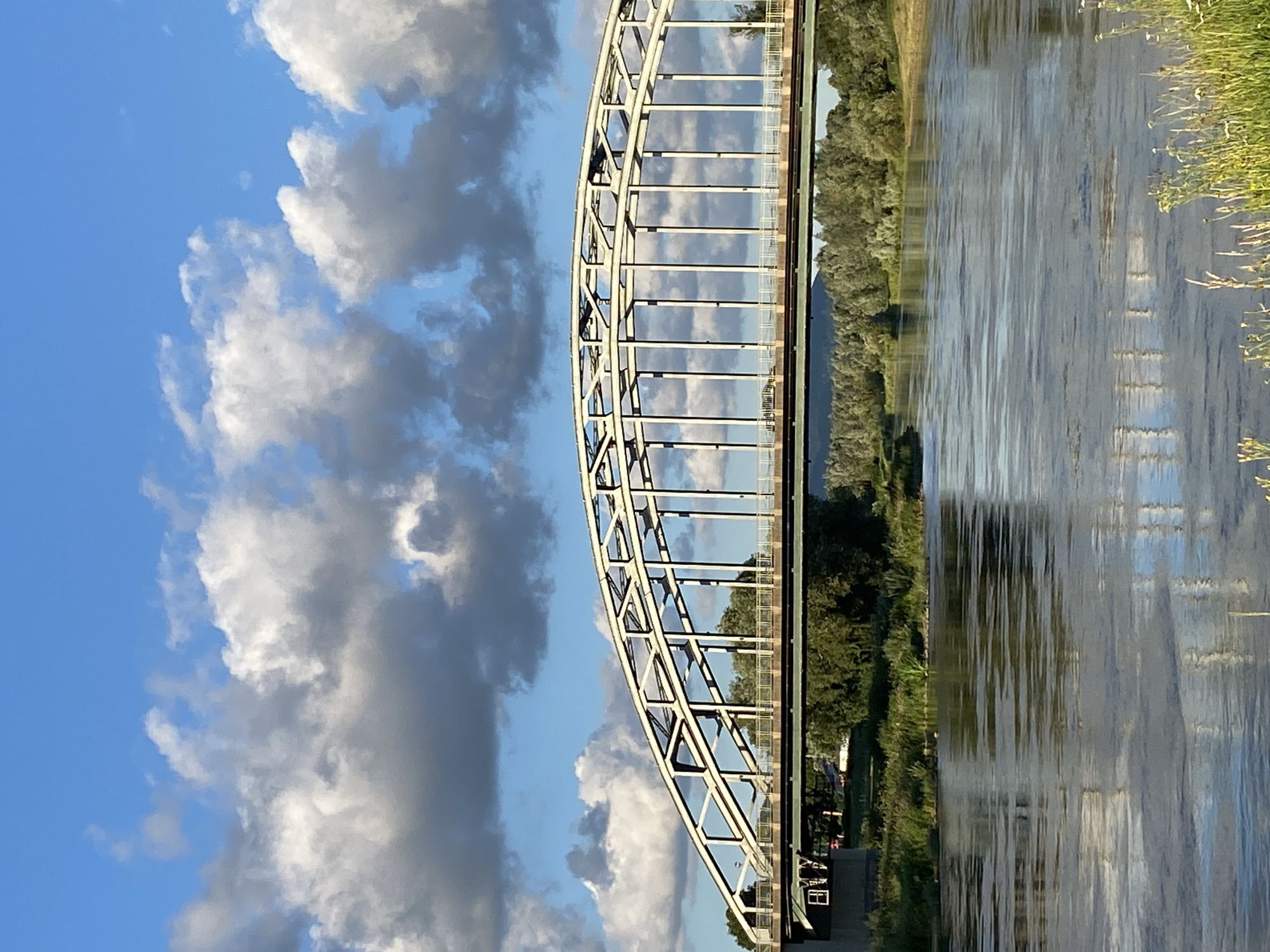 The image shows a large steel arch bridge spanning a wide river. The bridge is light-colored and has a distinctive lattice structure supporting the roadway. Vertical supports connect the arch to the road deck. The river is calm, reflecting the sky and the bridge. Along the riverbanks, lush green trees and vegetation are visible. The sky is blue with fluffy white and gray clouds. In the background, a distant treeline suggests a rural or suburban setting. The overall scene depicts a sunny day with dramatic cloud formations. This bridge is the Weserrenaissance Bridge (Weserrenaissancebrücke) in Rinteln, Germany.