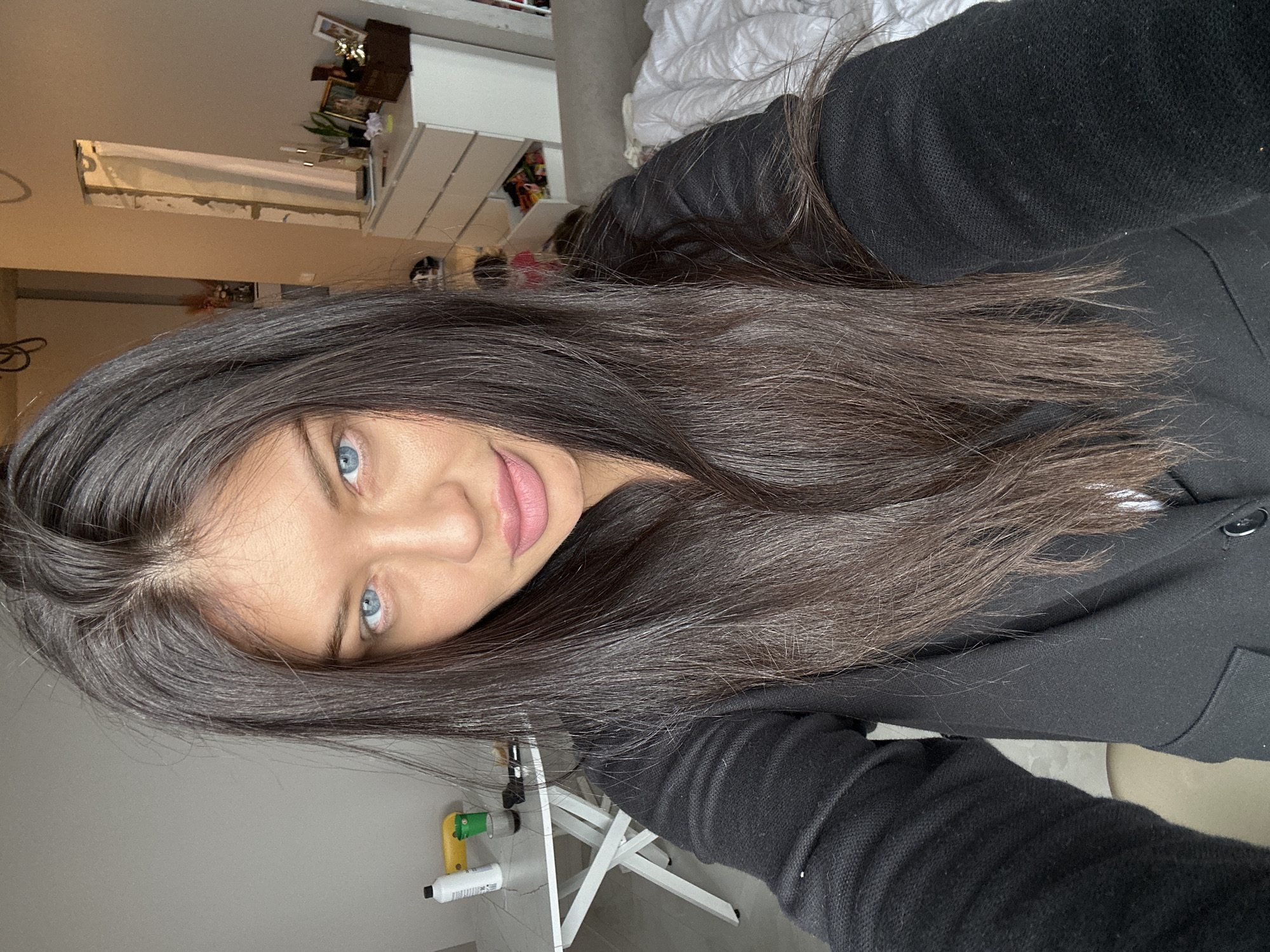 The image shows a woman with long, dark hair and blue eyes. She is wearing a black jacket. The background includes a white dresser, a bed with white bedding, and a white folding table with cleaning supplies. It is impossible to ascertain the location as Khimki, Russia based on the provided information.