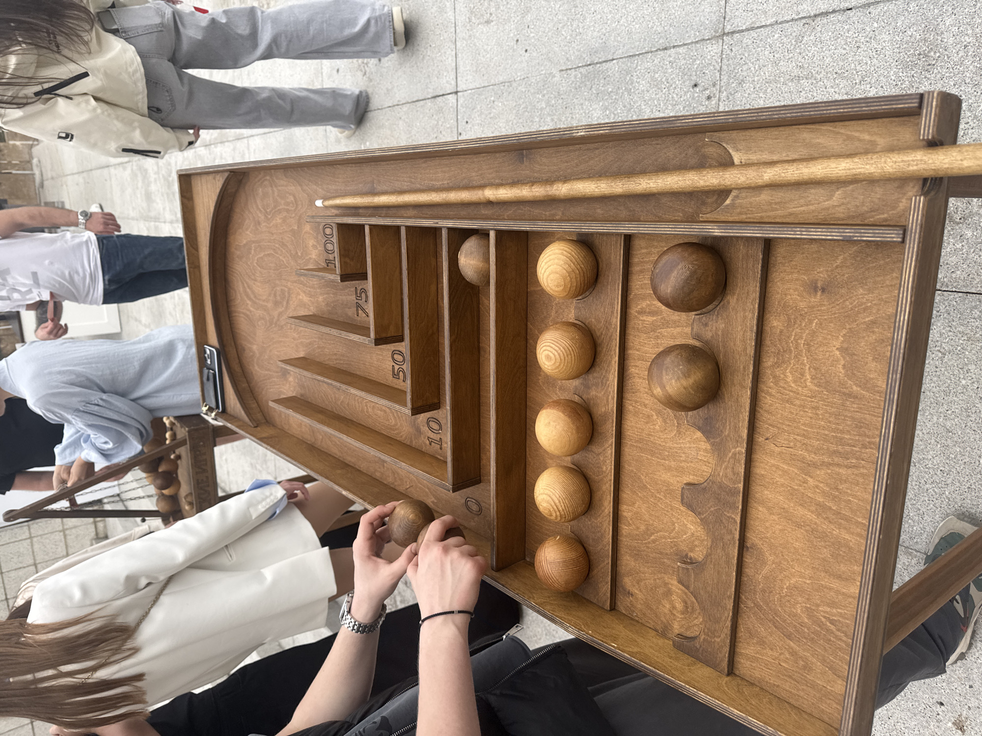 The image shows a wooden billiards-like game on a table with a cue stick. A person is holding a wooden ball in their hands, appearing to be playing the game. To the left, there are several people standing around. The game has score slots marked with numbers 10, 50, 75, and 100. The location is Sochi, Russia.