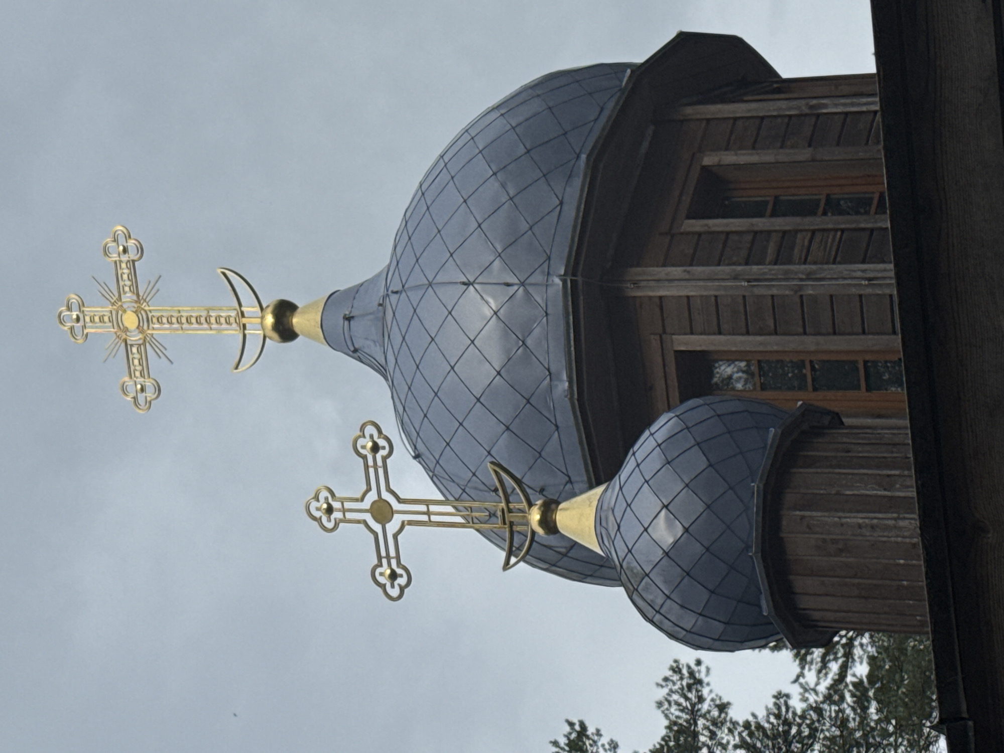 This is a close-up, eye-level shot of the tops of two onion domes of a wooden church, located in Nenyamyaki, Russia. The domes are covered in dark gray, diamond-shaped tiles. Each dome is topped with a golden cross. The larger dome is behind and to the right of the smaller dome. The wooden structure of the church is visible below the domes, with horizontal planks and vertical supports. Two windows with dark frames are visible in the larger dome's structure. A few evergreen trees are visible on the left side of the image, against a cloudy, overcast sky.