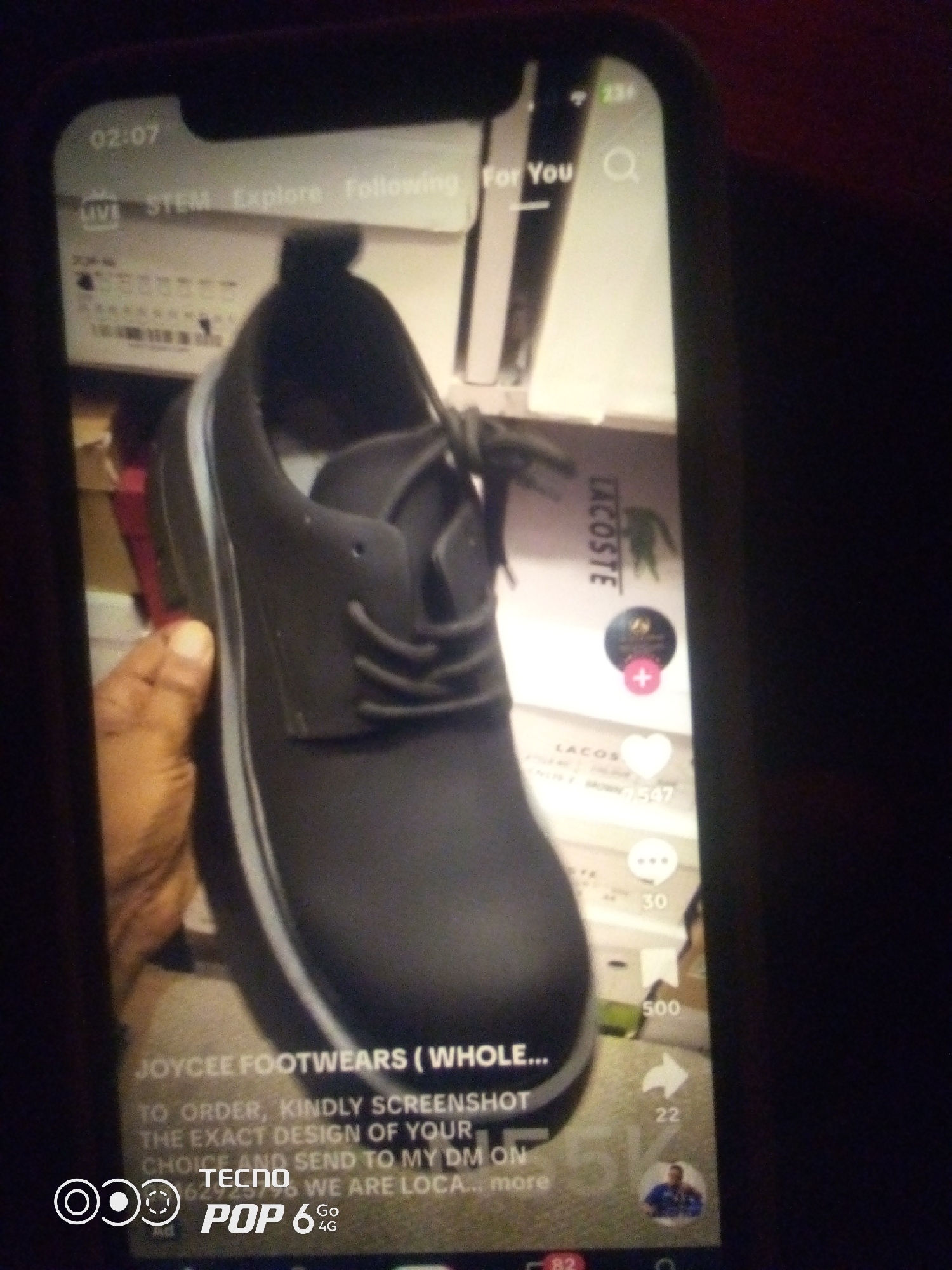 The image shows a close-up of a person's hand holding a dark-colored shoe, possibly a loafer or a casual shoe, on a smartphone screen. The shoe appears to be made of a matte material and has laces. The smartphone screen displays a social media interface, with text and icons suggesting a product listing or advertisement for "JOYCEE FOOTWEARS". The text also includes instructions to screenshot and send to a direct message for ordering. Other elements on the screen include what appears to be a brand logo (possibly Lacoste) and engagement metrics like likes and comments. The background of the shoe on the screen is a stack of boxes, some of which have branding visible. The phone itself is a TECNO POP 6 4G. The image is taken in a dimly lit environment. The specific location in Howayi, Nigeria, cannot be determined from the image content.