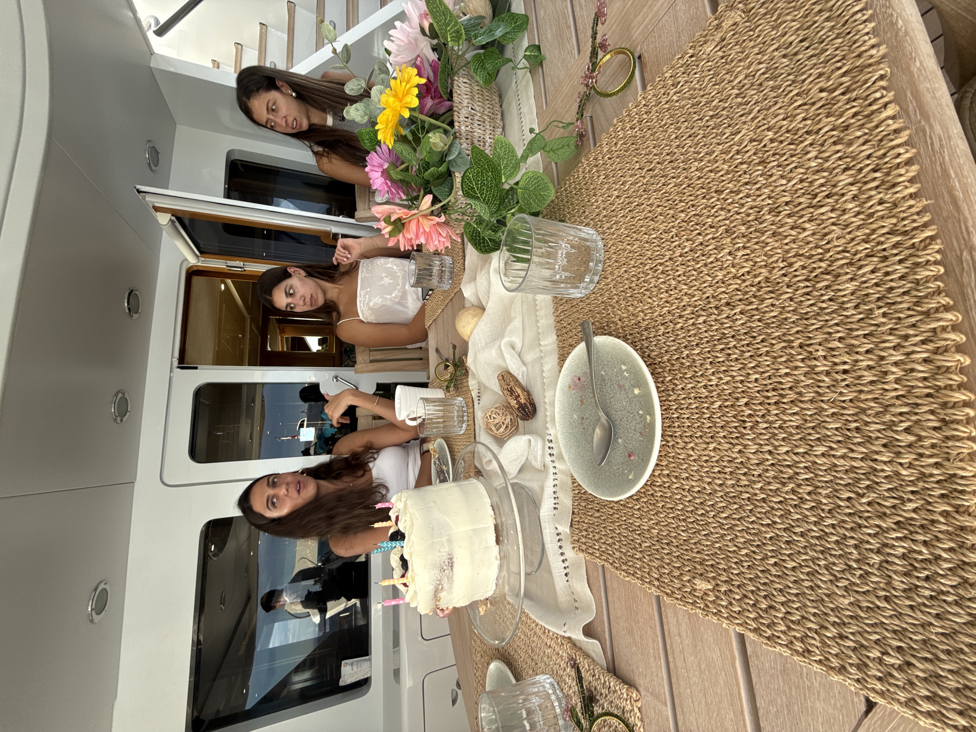 The image shows three women positioned on a boat. The boat's windows reveal scenes of La Paz, Mexico, including other vessels and reflective surfaces. A table is set up with a natural fiber runner. On the table, there is a cake on a glass stand with lit candles, a plate, and a spoon. Flowers are arranged in a vase and several glasses are present. Two of the women are looking toward the cake, while the third one is partly visible above them.