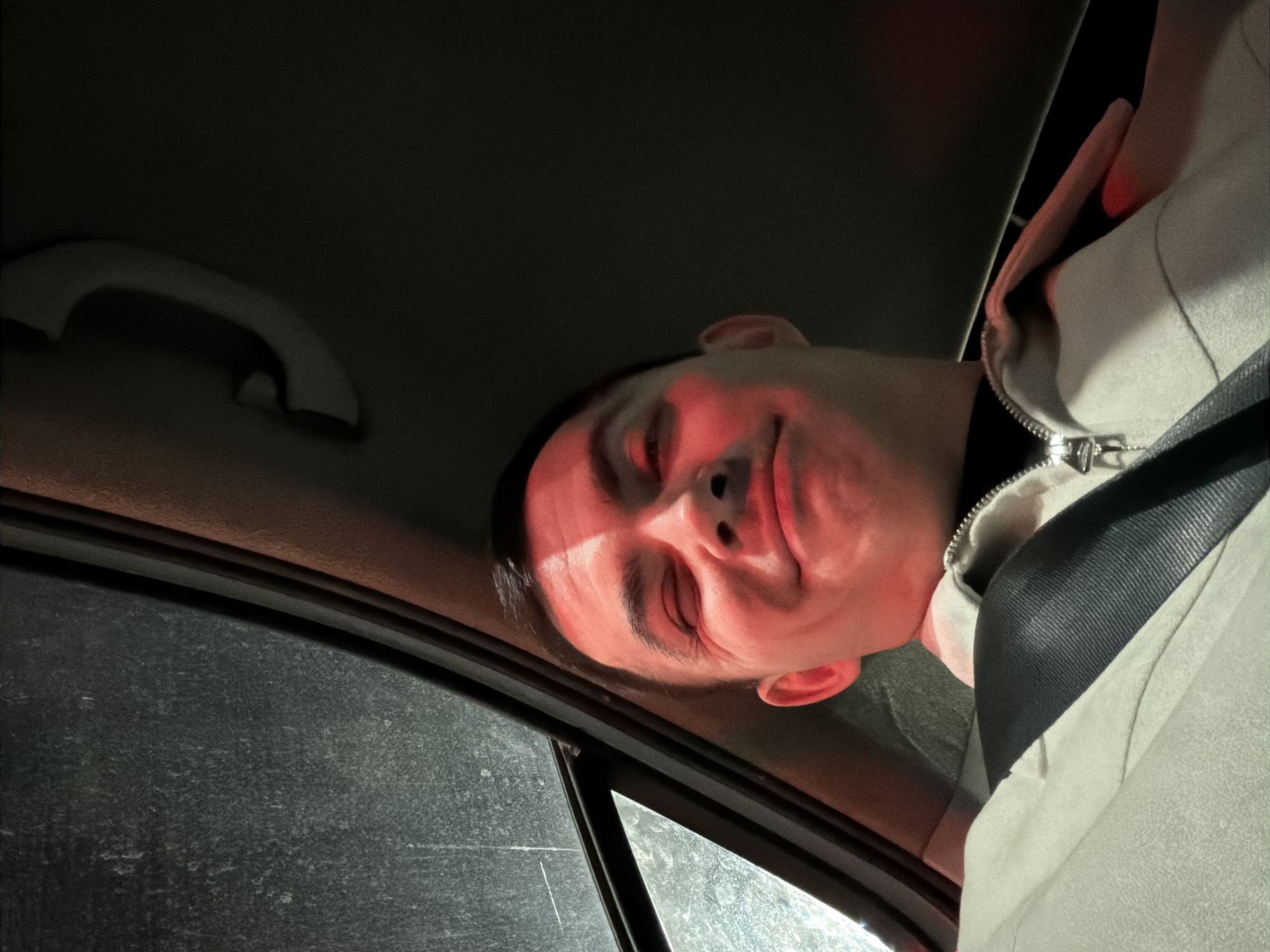 The image shows a person inside a vehicle, likely a car, with the camera positioned at an unusual angle. The person, possibly male, is wearing a seatbelt and a light-colored jacket with a zipper. The interior of the car is visible, including a grab handle and a window with grime. Red light illuminates the person's face. The location is not discernible from the provided image. There is no evidence to suggest this image was taken in the city of Izhevsk, Russia.