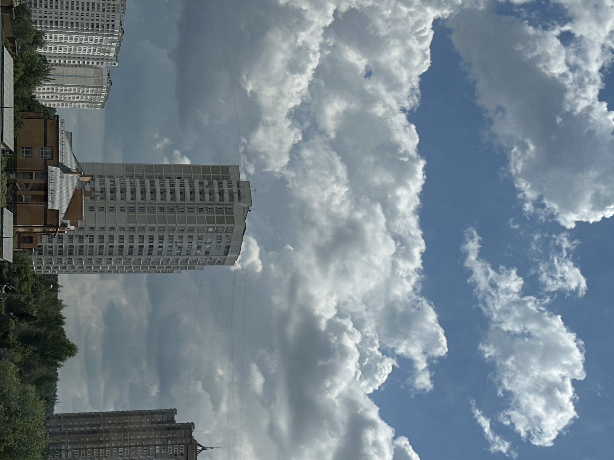 A high-angle outdoor shot depicts a cloudy sky over the city of Podolsk, Russia. Cumulus clouds, ranging in color from white to dark gray, dominate the sky, obscuring parts of the buildings below. Three residential high-rise buildings are visible. The tallest, in the center, is a light-colored, multi-story building with numerous balconies. To the left, two shorter, lighter-colored buildings with many windows are visible. To the right, another tall, dark-colored building with a unique top is partially visible. In the foreground, a low-rise brick building with a gray roof sits in front of the trees. The scene is dominated by the sky and buildings, with no people or vehicles in view.