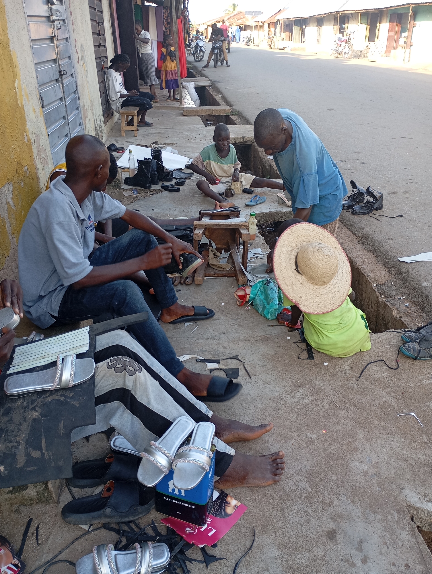In Shendam, Nigeria, two men and two children are engaged in shoe-making activities on a paved street in front of a row of shops. The men, one in a grey polo shirt and the other in a blue t-shirt, are seated on the ground. The man in the grey shirt is working on a sandal, holding it in his hands. The man in the blue t-shirt is leaning over, seemingly examining or working on something on the ground. A young boy in a green shirt and a straw hat sits nearby, also appearing to be involved in the shoe-making process. Another young boy in a t-shirt and shorts is seated to the left, looking towards the men. Various shoe-making materials, including leather scraps, boots, sandals, adhesive, and tools, are scattered around them on the ground. The environment suggests an outdoor workshop set up along a street, with a building wall and shop fronts visible in the background. The scene is illuminated by natural daylight.