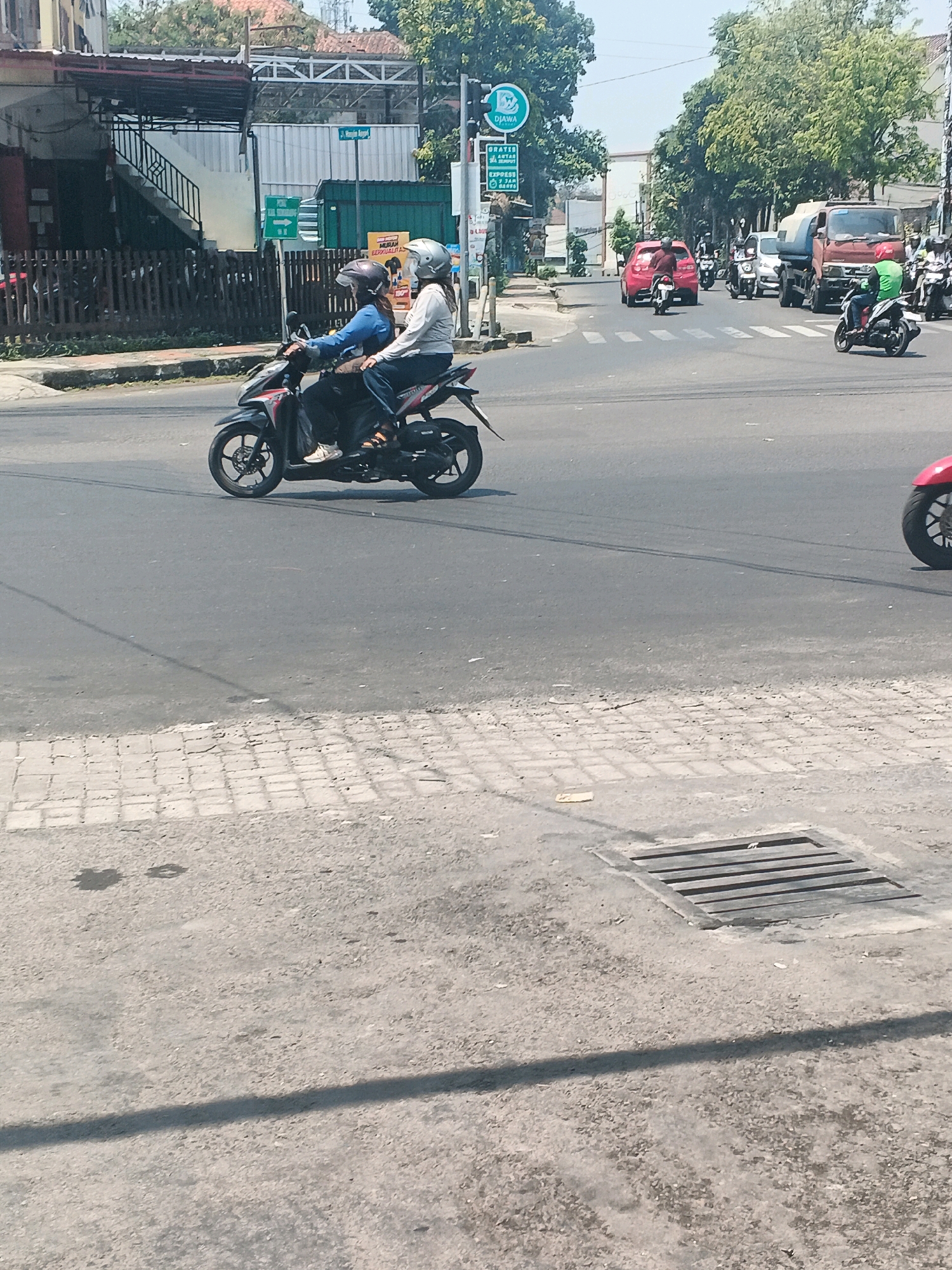 Two individuals, one driver and one passenger, are riding a black scooter in what appears to be a city intersection in Ungaran, Indonesia. The driver, wearing a blue jacket and a dark helmet, is steering the scooter. The passenger, wearing a light-colored jacket and a silver helmet, is seated behind the driver. They are proceeding forward on a paved road. In the background, there are other vehicles, including a red car, a truck, and several motorcycles, indicating a busy traffic environment. Buildings and trees line the sides of the road. The scene suggests a typical day with moderate traffic flow on a street in Ungaran.