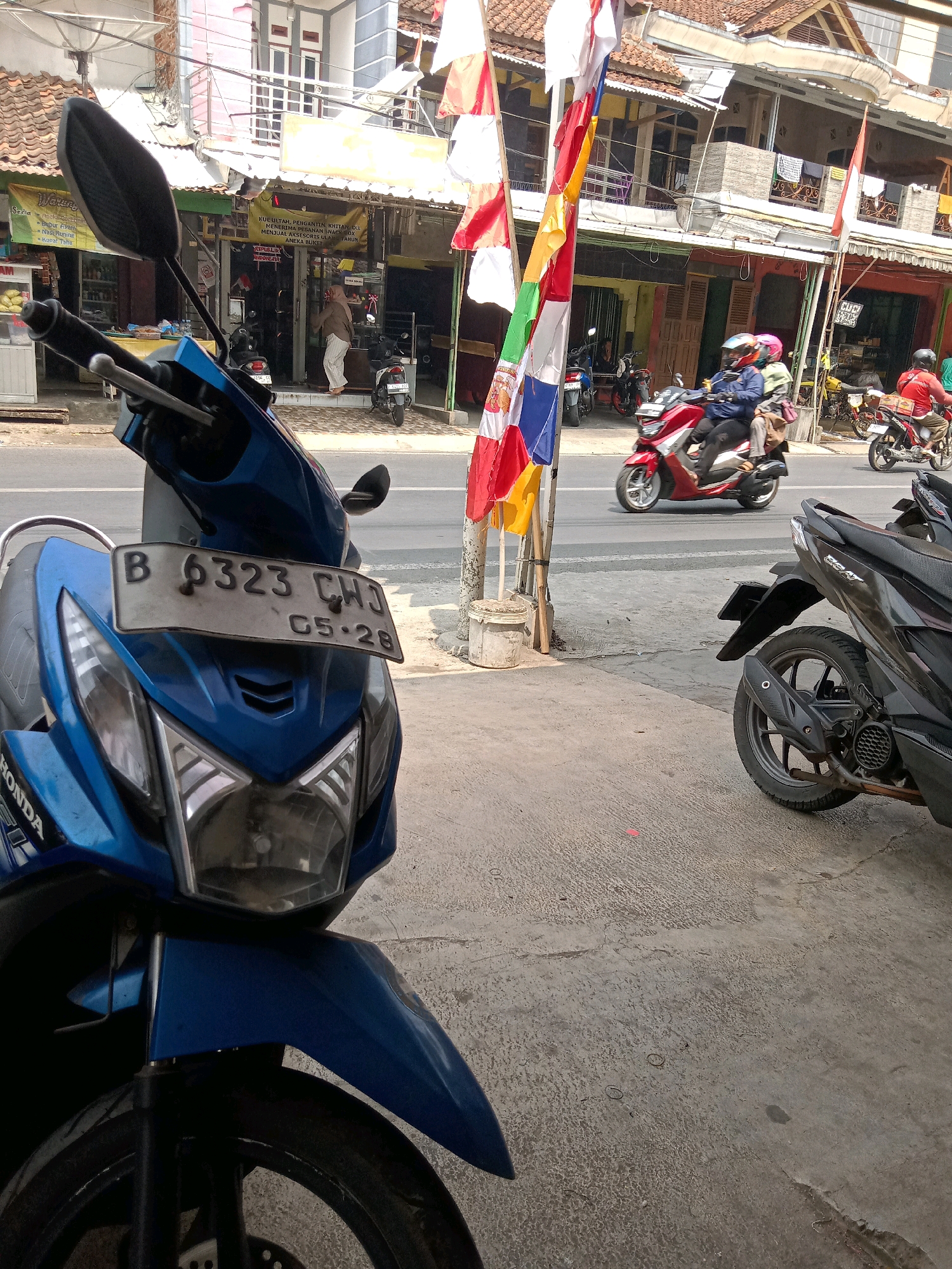 The image depicts a street scene in Sindangherang, Indonesia. In the foreground, a blue Honda scooter is parked, its front end and handlebars visible. The scooter has a license plate that reads "B 6323 CHU" and below it "05-28". Behind the blue scooter, a black scooter is also parked, only its rear wheel and fender are visible.
Across the street, there are several buildings with shops. Flags, predominantly red and white, are displayed vertically along the sidewalk in front of one of the shops. Two motorcycles are in motion on the street. One is a red scooter with two riders, and another is a white scooter further down the road. Pedestrians can be seen walking in front of the shops. The environment appears to be a commercial area with a paved road and sidewalk, suggesting a typical urban setting. The presence of Indonesian flags indicates a possible national holiday or celebration.