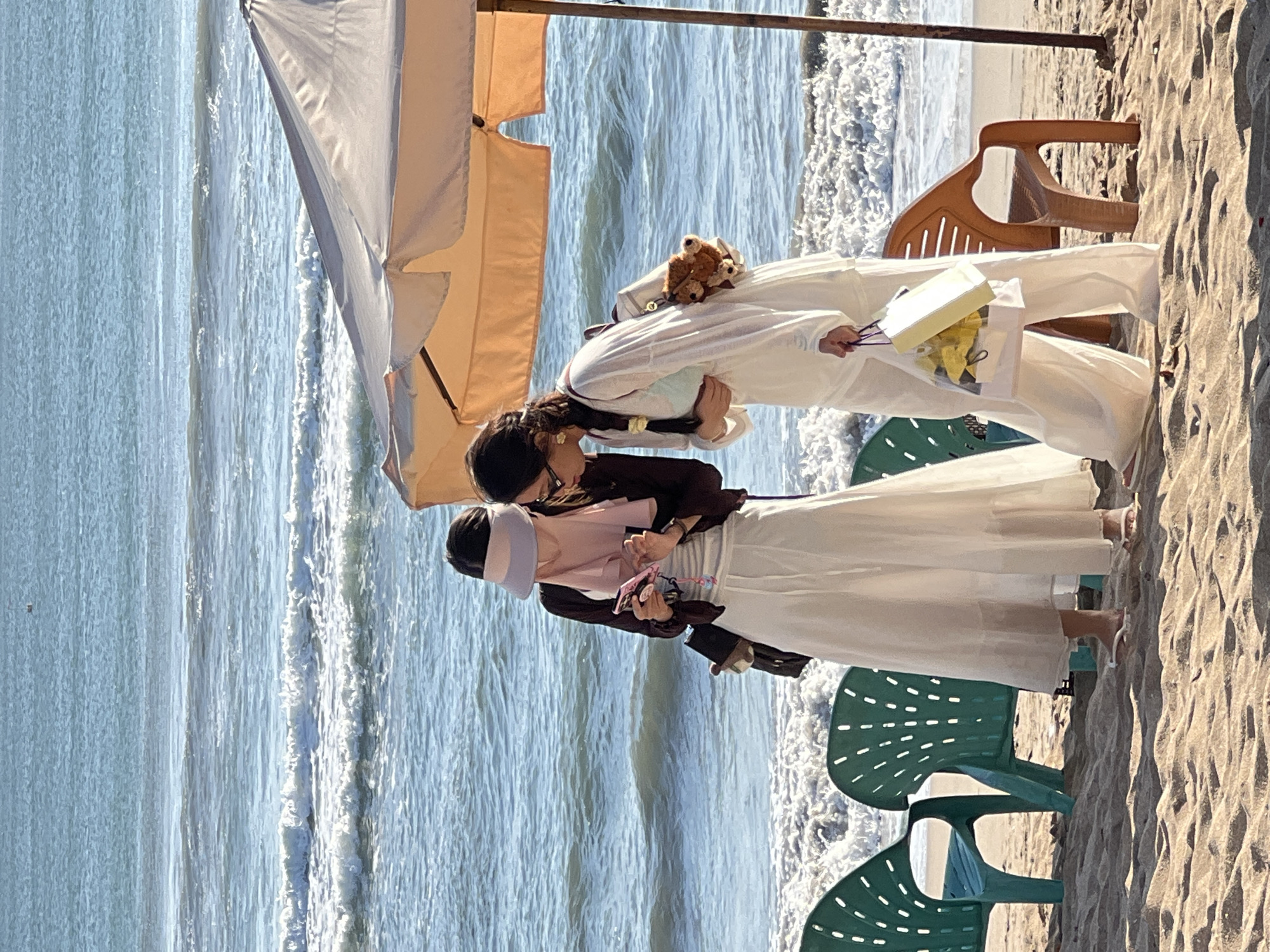 Two women are on a sandy beach in Kuta, Indonesia, near the ocean. The woman on the left, wearing a white skirt and a pink headscarf, is looking at her phone. The woman on the right, wearing white pants and a white long-sleeved shirt, is also looking at her phone and holding a shopping bag. A beach umbrella casts a shadow over them. Green plastic chairs are visible in the foreground, and the ocean waves are crashing on the shore behind them. The scene appears to be during daylight.