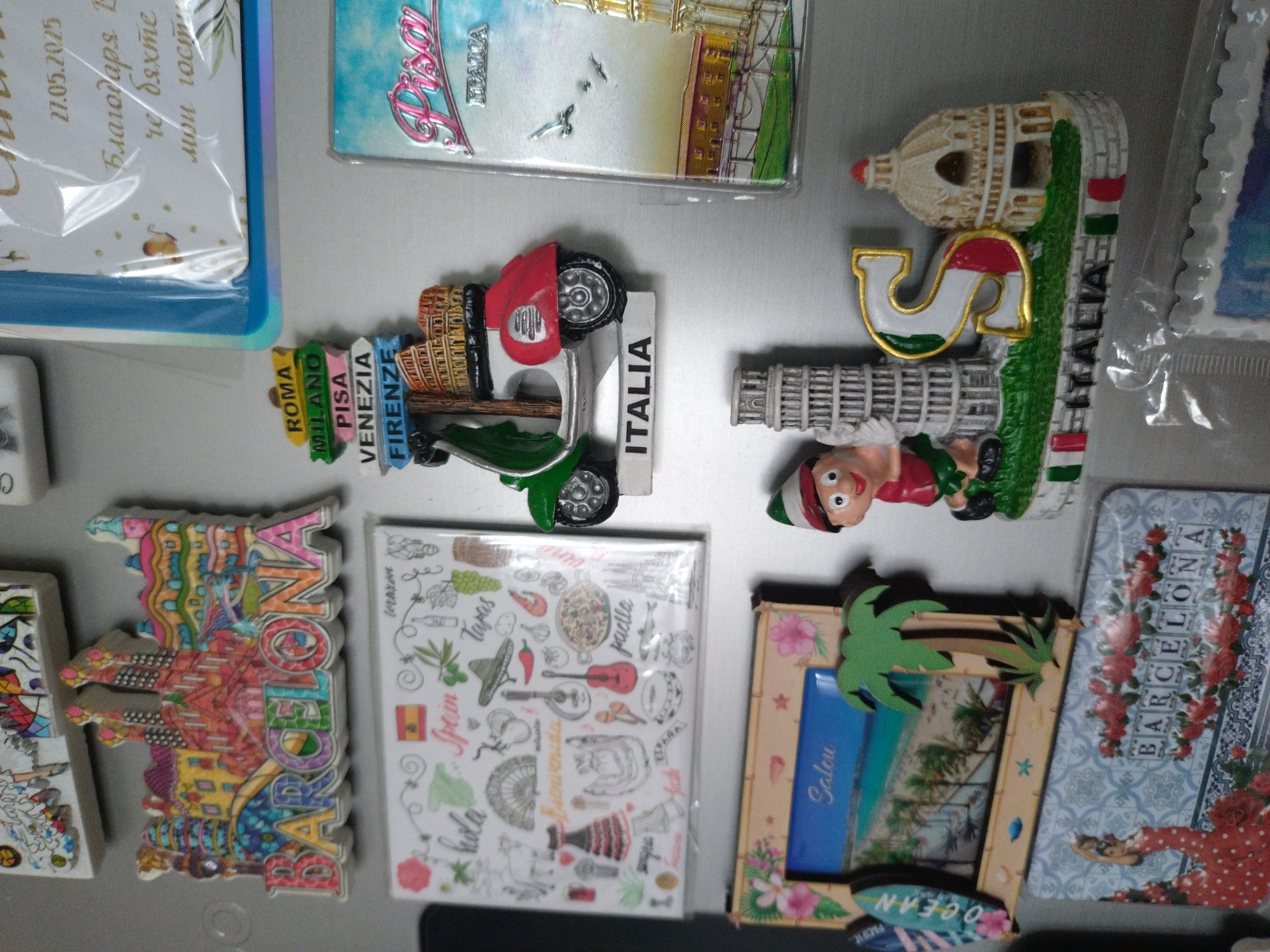 The image shows a collection of refrigerator magnets. The magnets feature various tourist destinations, including those in Italy (Rome, Milan, Pisa, Venice, Florence, and generally "Italia" depicted with a leaning tower and a chef), Spain (Barcelona and various Spanish cultural elements). One magnet appears to depict the city of Salou, while others have generic seaside or ocean themes. A date "27.05.2025" and text written in Cyrillic (likely Bulgarian) is also seen, suggesting the image could be from Bulgaria. The location of Stara Zagora is not explicitly represented; however, the Cyrillic text implies the possibility of Bulgarian origin.