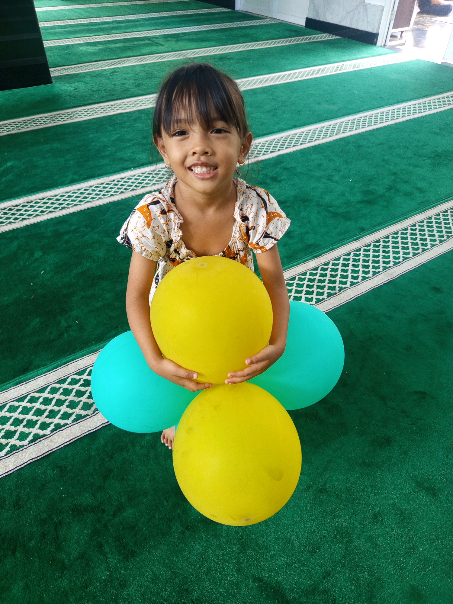 A young girl with black hair and a white and orange patterned top is smiling and holding two yellow balloons and one teal balloon in front of her. She is sitting on a green carpeted floor with white striped patterns. The setting appears to be a prayer room or a similar space with the characteristic patterned carpet. The girl's lower body and feet are visible, suggesting she is seated on the floor. The lighting is bright, coming from the right side of the frame. Based on the distinctive prayer carpet pattern, the location can be identified as a mosque or a prayer room in Bogor, Indonesia.