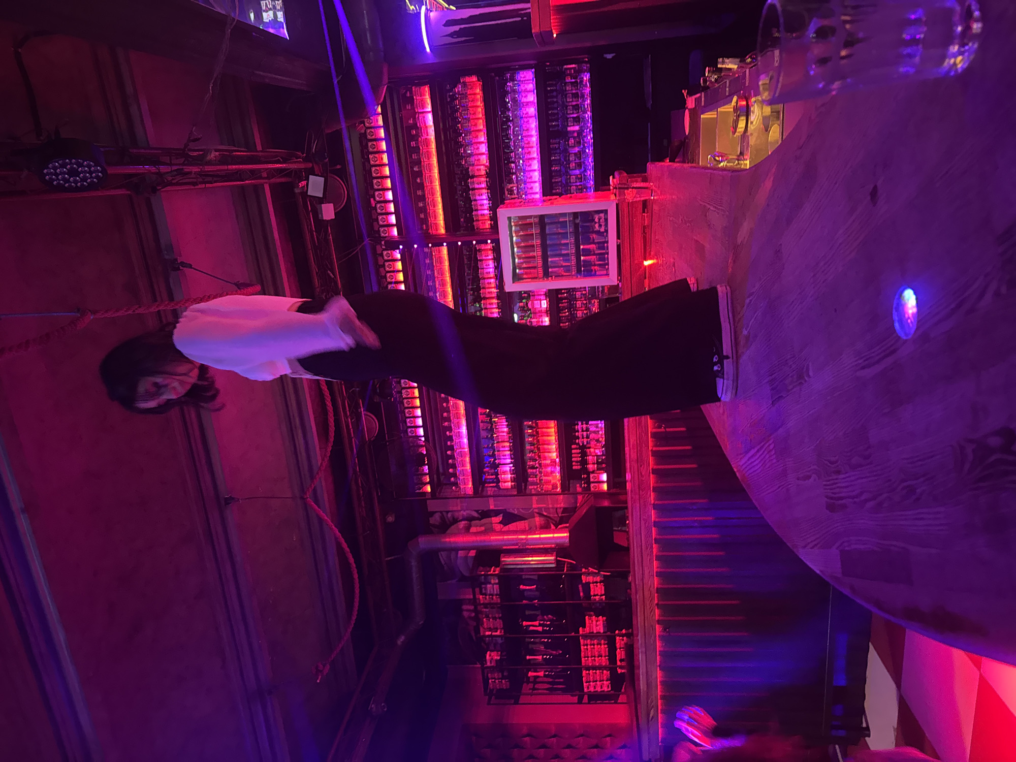 A young woman with dark hair, wearing a white crop top and black pants, stands on a wooden bar counter. She is captured in mid-movement, appearing to be dancing or posing. The scene is set in a dimly lit establishment, possibly a bar or nightclub, with vibrant pink and purple lighting casting a neon glow. Behind her, shelves are filled with numerous bottles of alcohol, illuminated by red and pink lights. A refrigerator stocked with red bull cans is also visible on one of the shelves. The bar counter she stands on is wooden and appears to be the central focus of the foreground. In the background, structural elements of the ceiling and lighting fixtures are visible. The overall atmosphere suggests a lively nighttime venue.