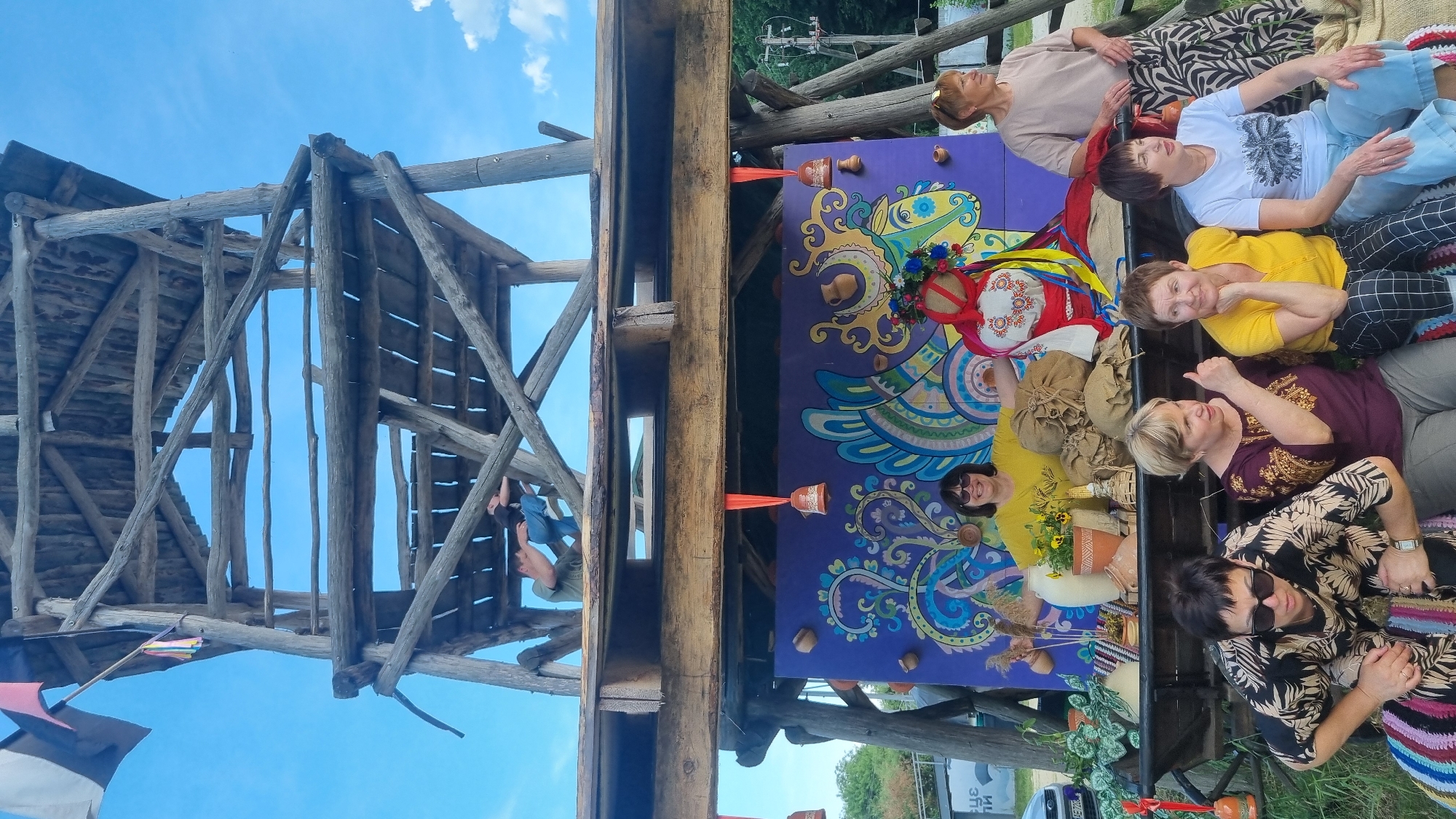 This image depicts an outdoor scene in Melnyky, Ukraine, featuring a multi-tiered wooden observation or decorative tower and a lower, decorated stage area, with a group of people gathered around it under a clear blue sky with sparse clouds.
The upper portion of the image shows a tall, rustic wooden tower constructed from logs and planks. On an upper platform of this tower, one individual wearing a green shirt and blue jeans is visible, appearing to be lying or climbing. Flags (white, black, orange/red) are attached to a pole extending from the very top left of the tower.
Below the tower, a wider wooden structure supports a vibrant purple backdrop adorned with colorful folk art patterns, including a large, stylized bird or mythical creature design. Small clay pots are attached to the backdrop, and two orange ribbons suspend bell-shaped objects. In front of this backdrop, on a wooden surface that functions as a display, are various traditional items: a large ceramic jug with flowers, smaller pottery vessels, bundles of burlap, woven artifacts, dried plants, and a large Ukrainian folk doll (motanka) with a floral headpiece and colorful ribbons.
Seven women are present in the foreground and mid-ground. One woman in a yellow shirt and sunglasses stands behind the display, smiling towards the camera with her arms slightly spread. To her right, another woman in a light tan top and a patterned skirt stands, looking to the right. In the immediate foreground, five women are seated on a colorful striped mat. From left to right: one woman in a black and tan patterned top and sunglasses looks down; next, a woman in a purple embroidered top gives a thumbs-up gesture; followed by a woman in a yellow t-shirt and checkered pants, resting her chin on her hand; then a woman in a white graphic t-shirt and blue jeans, looking slightly to the right; and finally, a partially visible woman behind her.
The ground is covered with green grass, and lush green foliage is visible in the background. A white vehicle is partially visible on the far left.