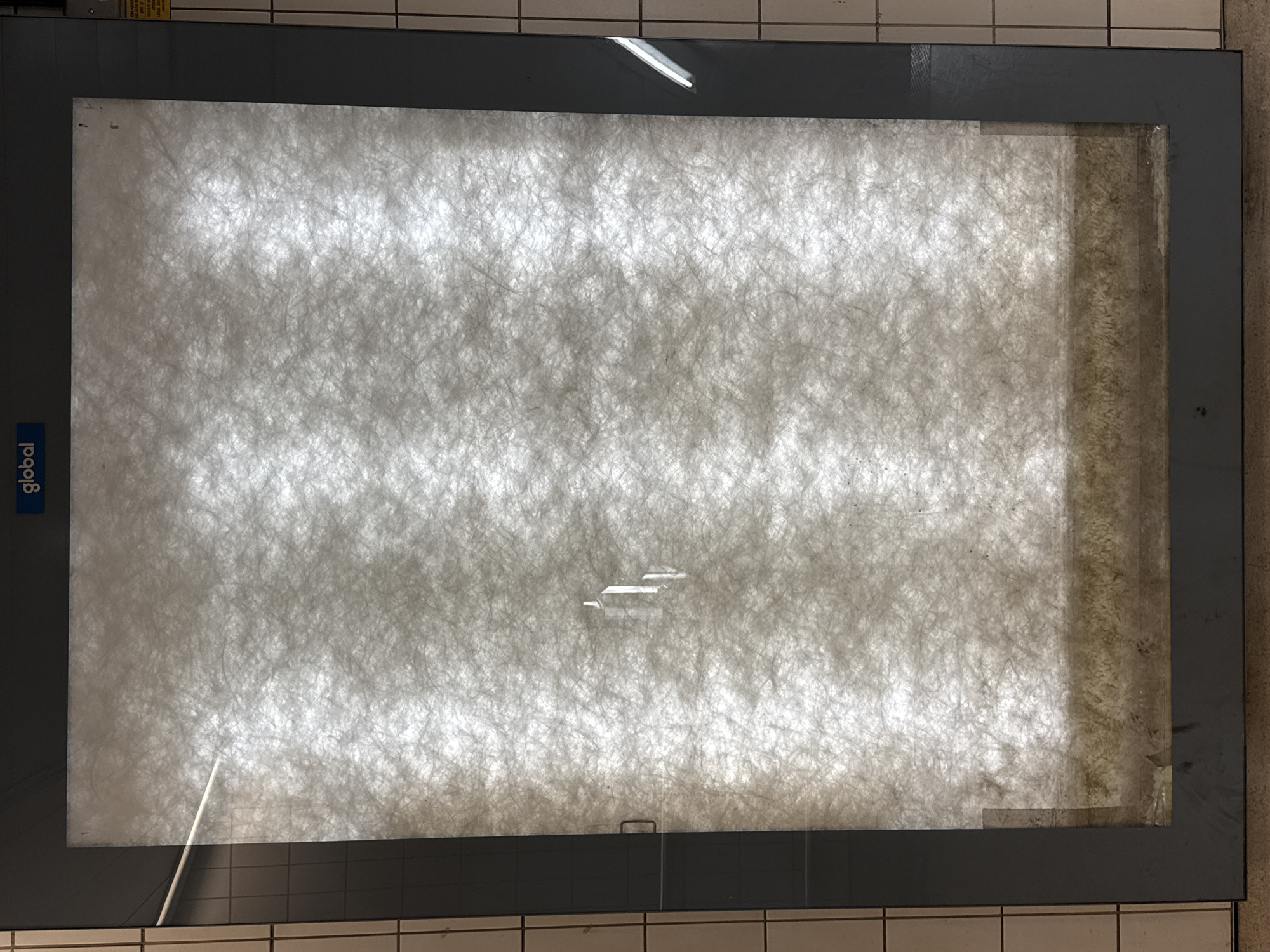 A gray-framed display case is mounted on a tiled wall. The case contains what appears to be a dirty air filter. The word "global" is written vertically in blue on the left side of the frame. The display case is located in a public area with tiled walls and floors. The location is London, United Kingdom.