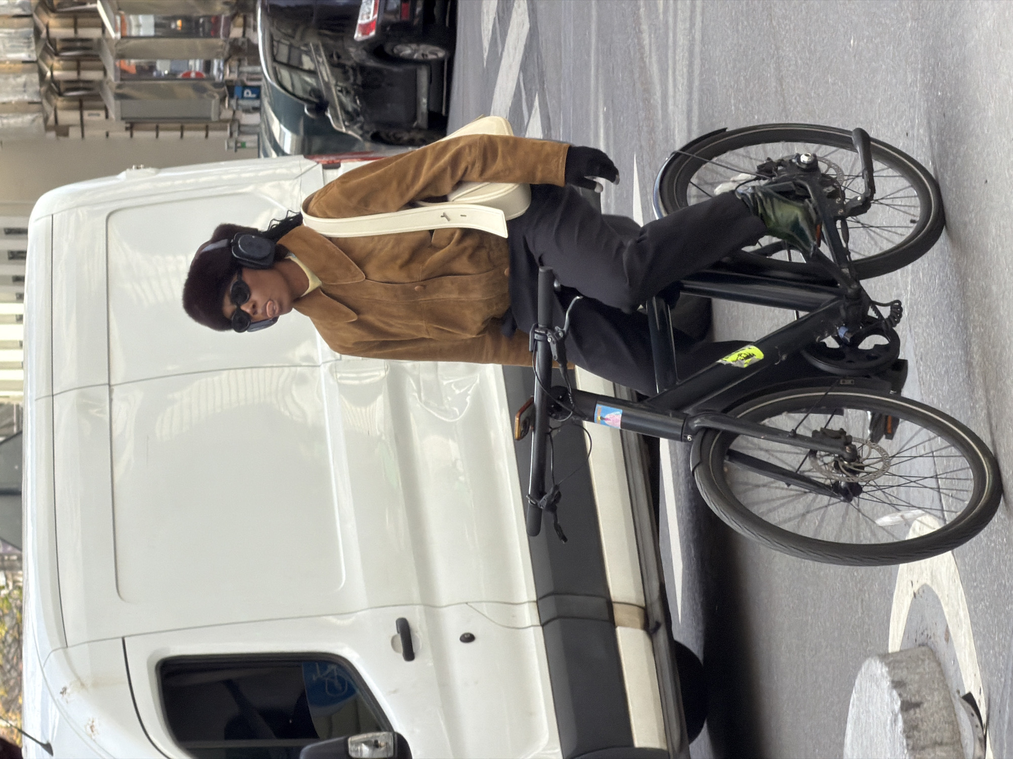 The image shows a person on a black bicycle on a street adjacent to a large white van. The person is wearing a brown jacket, black pants, gloves, headphones, and sunglasses. A white bag is slung over their shoulder. The background includes a blurred view of other vehicles and buildings, suggesting a city street. The presence of a van and general urban setting suggest the scene is potentially located within a city. There is no specific identifiable marker in the photo itself that confirms its location as Paris, France. Without more detailed information, I cannot conclusively confirm the location as Paris.