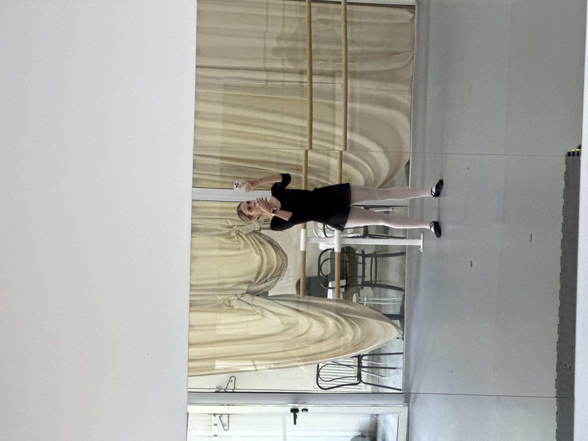A young woman, likely a dancer or ballet student, is captured taking a mirror selfie in what appears to be a dance studio. She is standing upright, holding a white smartphone in her right hand, angled towards the large mirror she is posing in front of. Her left hand is raised slightly, and she is wearing a black leotard or dress, white tights, and black ballet slippers. A watch is visible on her left wrist.
The setting is characterized by a full-length mirror reflecting the studio space. Behind the woman's reflection, multiple wooden ballet barres are visible, running horizontally across the wall. Behind the barres, light-colored, possibly beige or cream, curtains are drawn, creating a draped background. Several chairs, some dark-colored and others lighter, can be seen reflected behind the curtains. A door frame or window frame is also visible at the bottom edge of the mirror reflection.
Outside the mirror's frame, a plain white wall occupies the left side of the image, while on the right, a grey floor made of interlocking exercise mats is partially visible. The lighting is bright and even, suggesting it is daytime. There is no visible text in the image. The location context for this scene is Tbilisi, Georgia.
