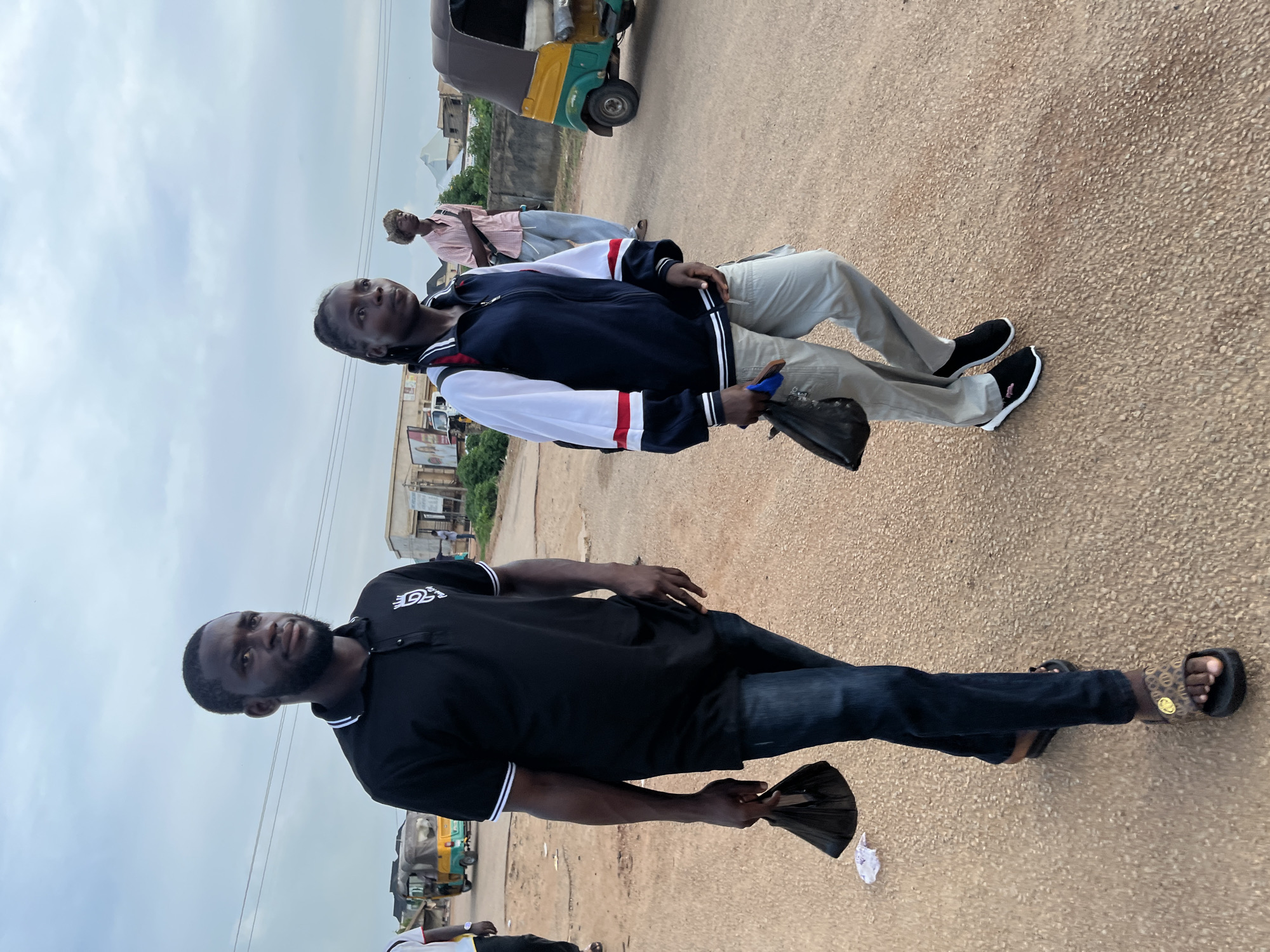 This image captures a daytime street scene in Rafin Par, Nigeria, under an overcast sky. The foreground features a man standing on a light brown road, dressed in a black polo shirt with white collar and sleeve stripes, dark blue jeans, and brown patterned slides with a circular logo. He has a beard, looks towards the viewer, and holds a crumpled black plastic bag. A white logo, appearing to be "DS" with stylized text, is visible on his shirt's left chest.
To his right, a younger person wears a navy blue and white jacket with red stripes, light grey trousers, and black slip-on shoes featuring white soles and a small pink polo pony logo. This individual looks slightly right of the camera, holding a dark object. Further in the background, another person in a light pink shirt and blue trousers is partially visible.
The setting includes simple buildings and sparse vegetation along the roadside. Overhead electrical wires are visible. Two auto-rickshaws, common in Nigeria, are seen; one behind the person in the jacket and another further back to the left. The scene depicts typical street activity in a semi-urban environment.