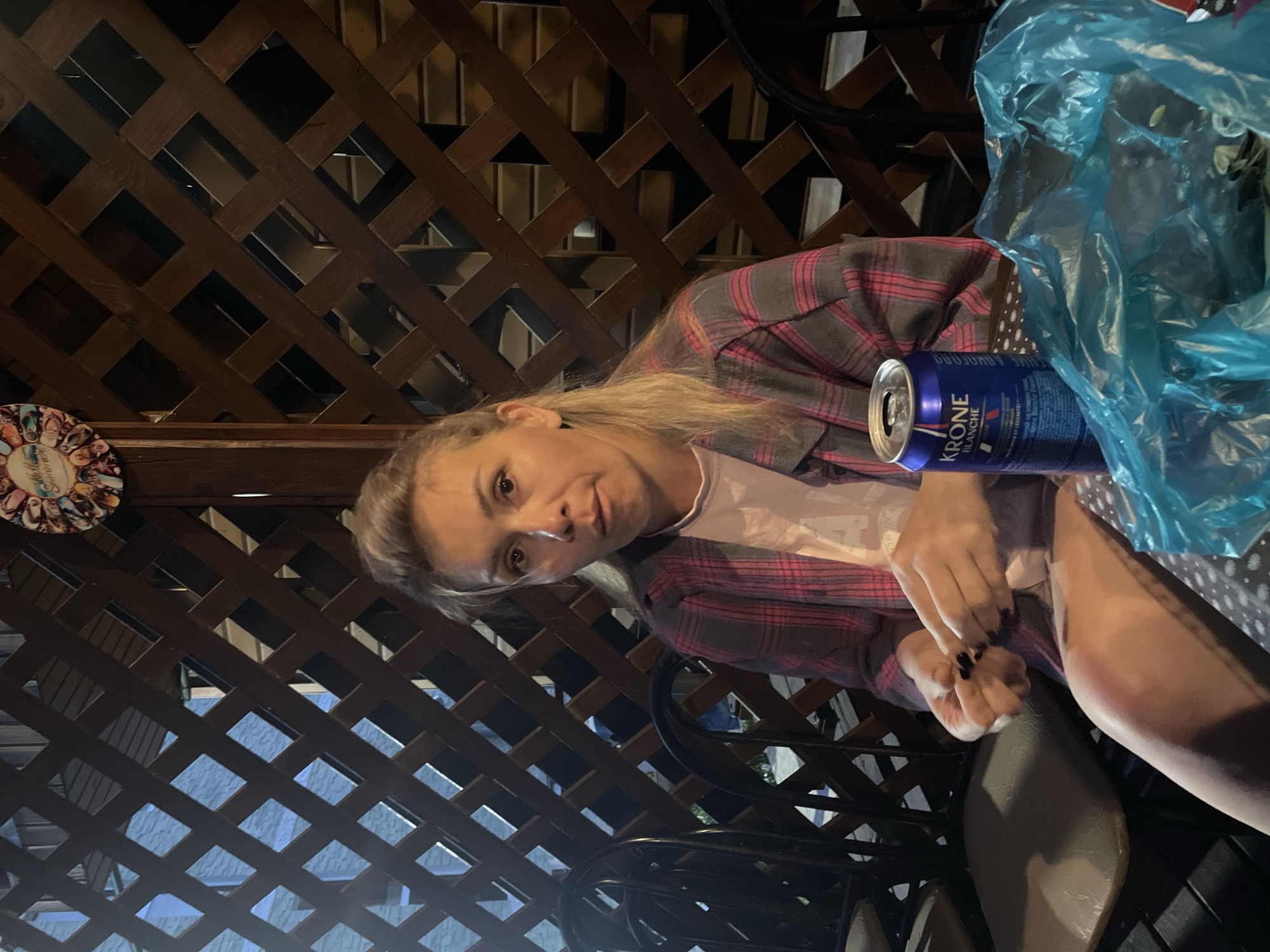 A woman with blonde hair and fair skin sits in front of a wooden lattice wall. She is wearing a pink and gray plaid shirt over a light pink t-shirt and a polka dot skirt. Her right hand is holding something small, and her left hand is raised, with her fingers extended. A blue can with "KRONE BLANCHE" printed on it sits on a surface in front of her, next to a crumpled blue plastic bag. Chairs are visible to her left and right. The setting appears to be outdoors, possibly a patio or gazebo, with a decorative plate hanging on the wall behind her. The lighting is dim.