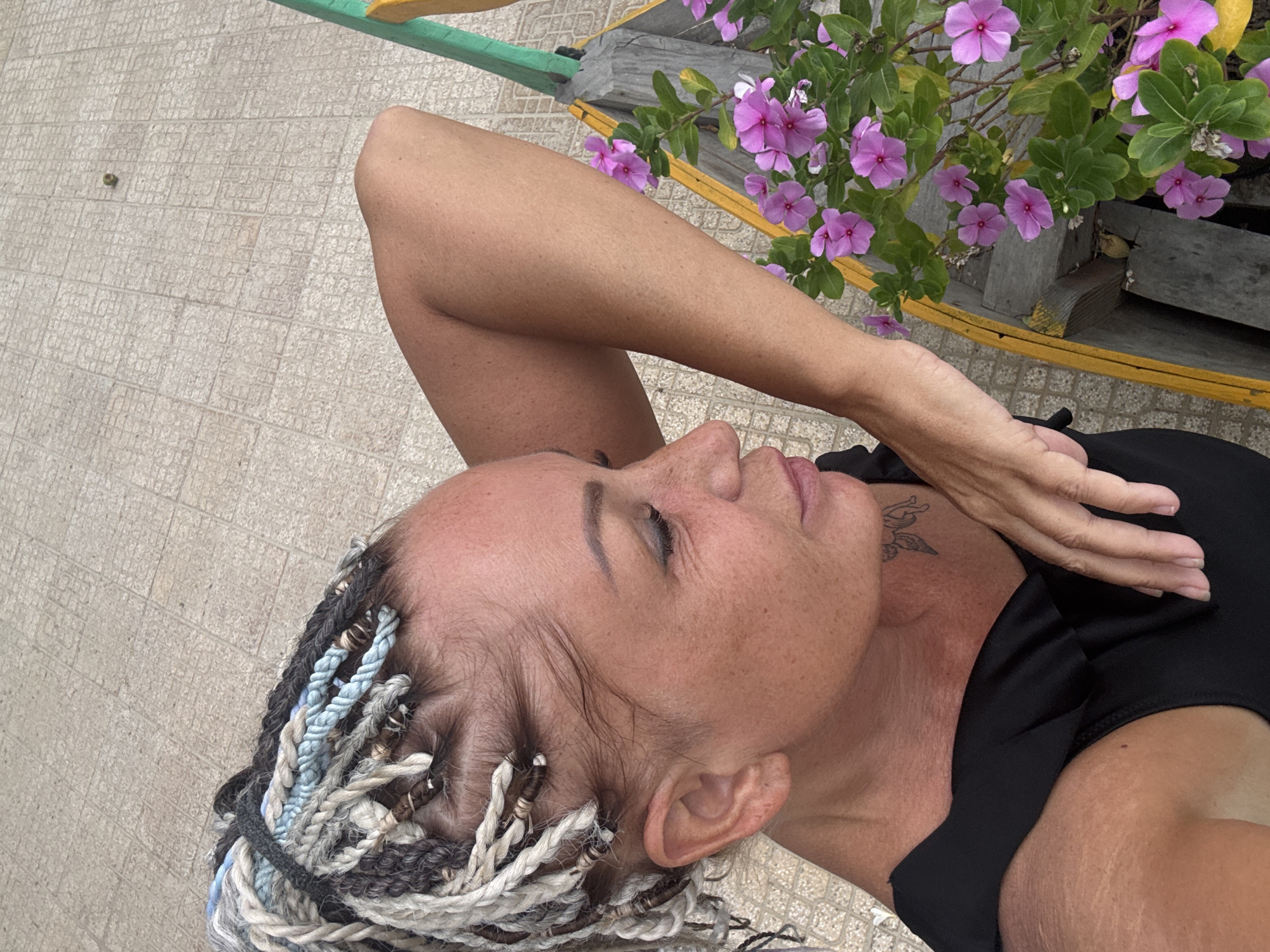 An overhead shot features a woman in a black garment, possibly swimwear, reclining near a flower planter with pink blossoms. She has light skin, a visible tattoo on her neck, and her hair is styled in braids with various colors including white, blue, and brown. Some braids have metal rings. She is lying on a tiled surface. Green and yellow painted wood trim appears in the background near the flowers, suggesting a built structure. While the prompt asks for the city of Đà Nẵng, Vietnam, the image does not contain definitive markers confirming that specific location.