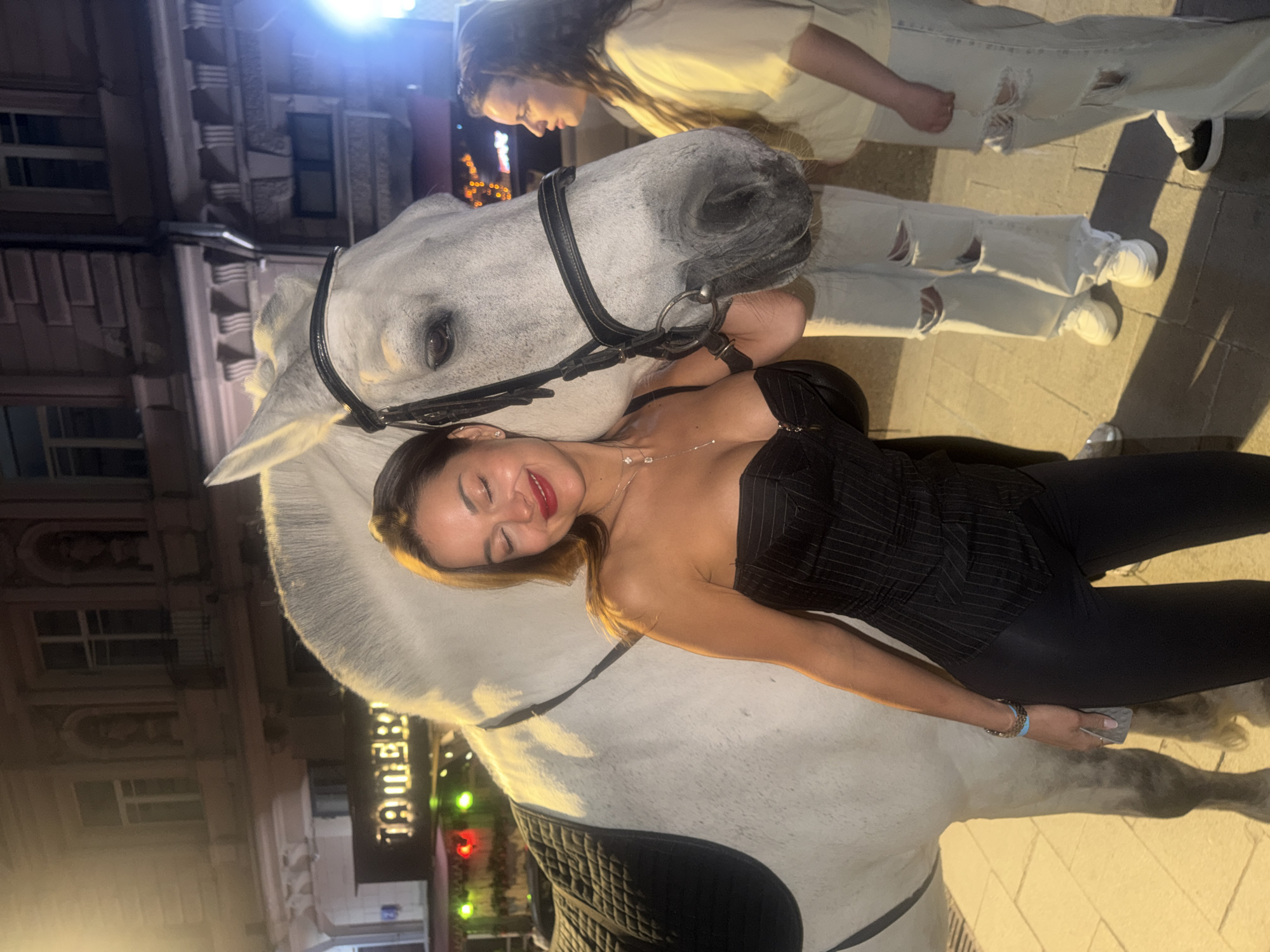 The image displays a nighttime urban scene in Moscow, Russia, featuring a large white horse and three individuals.
In the foreground, a Caucasian woman, appearing to be in her late 20s or early 30s, is positioned closely with the horse. She has long dark brown hair, red lipstick, and is smiling with her eyes closed, resting her head against the horse's neck. She wears a black strapless corset-style top with subtle vertical pinstripes, black tight-fitting pants, a delicate silver necklace, a small earring in her left ear, and a blue/brown bracelet on her right wrist. Her left arm is around the horse's neck, and her right hand holds a small object near her hip.
The horse is a large white or light grey dapple equine, equipped with a black leather bridle and a black quilted saddle. It stands placidly, with its head turned slightly towards the woman.
To the right, in the mid-ground, a second Caucasian woman, also appearing to be in her late 20s or early 30s, stands wearing a light yellow oversized t-shirt and light-wash ripped jeans with white sneakers. Her long dark hair is down, and she observes the interaction between the first woman and the horse. Partially visible between the horse and the second woman are the lower legs and feet of a third individual, dressed in similar light-colored ripped jeans and white sneakers.
The background features a multi-story building with classical architectural elements, including ornate window frames. A brightly lit sign displaying "ТАВЕРНА" (Tavern) in Cyrillic characters is visible on the building. The ground is paved, and the scene is illuminated by ambient artificial lighting from street fixtures and building lights, with a strong light source casting a glare from the right side of the frame.