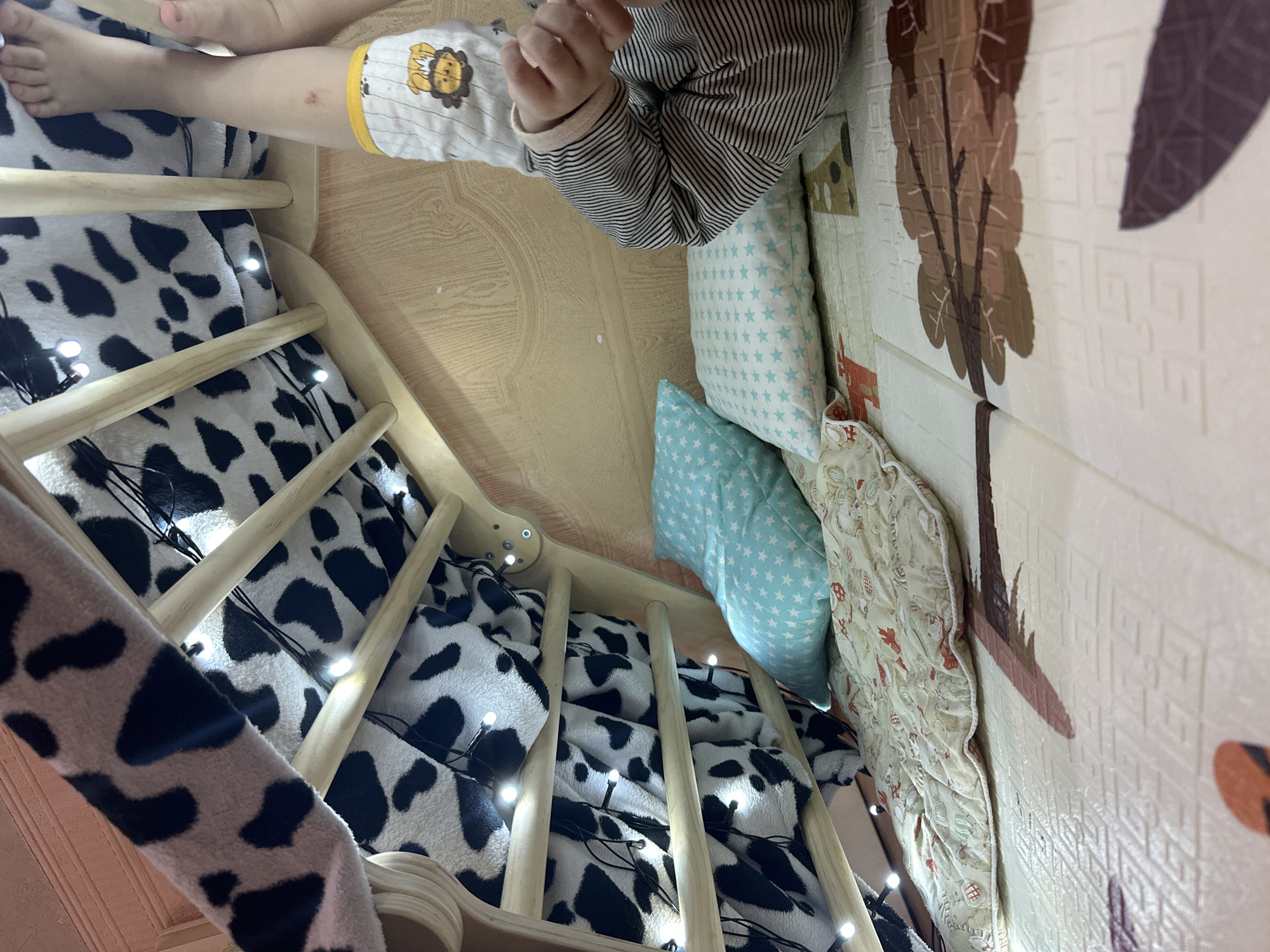 The image depicts an indoor scene, likely a child's play area. A wooden climbing structure is visible, draped with a black and white cow-print fabric and adorned with fairy lights. Below the structure, two blue star-patterned pillows and a cream-colored quilted mat with red and beige patterns are placed on a large, light-colored play mat featuring tree designs. The lower right corner of the image shows a child's legs and feet. The child is wearing striped pants and a diaper with a cartoon lion print. A small reddish mark is visible on the child's leg. The wall in the background is made of light-colored wood. There is no information in the image to suggest the location is Vladivostok, Russia.