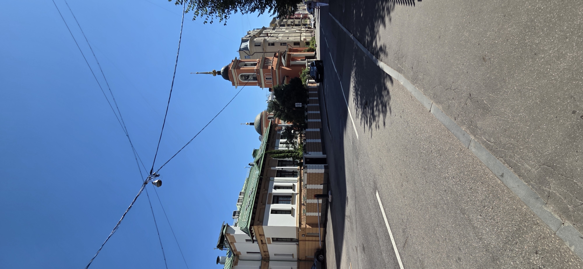 This is a street-level, vertical, eye-level shot of a street in Moscow, Russia, on a clear, sunny day. In the foreground, there is an asphalt road with white lane markings. The right side of the road has a sidewalk with a grey curb. Trees cast shadows on the road from the right.
On the left side of the road is a light-colored, multi-story building with a green roof and decorative cream-colored accents. It has several dark windows. Adjacent to it, and slightly behind, is an orange-colored church with a bell tower topped with a golden dome. The church has arched windows and a bell visible in the tower. Behind these buildings, other residential and commercial buildings are visible, extending down the street.
There are power lines suspended across the top of the frame, with a street lamp visible. A dark-colored car is parked on the right side of the street, and another dark-colored car is parked on the left side of the street. A person in light clothing is walking on the sidewalk on the far right side of the image. The overall environment appears to be a well-maintained urban street.