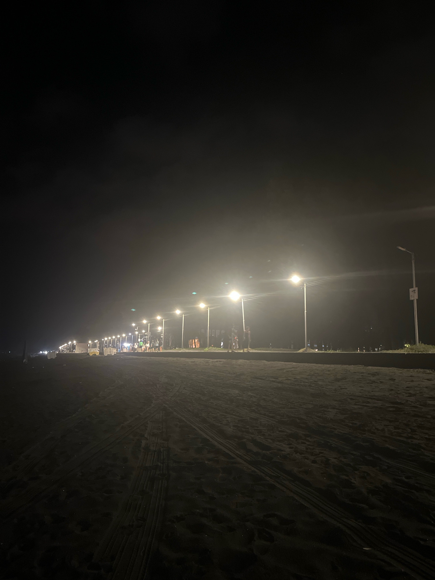 This is a nighttime photograph of the Ureki, Georgia beach. The scene is illuminated by a series of streetlights, casting a warm glow across the sand. The beach is mostly empty, with only a few scattered figures visible in the distance. Tire tracks are imprinted on the sand, suggesting that vehicles have recently traversed the area. In the background, the lights of the town are visible, creating a soft haze against the dark sky. The overall impression is one of tranquility and quiet, with the natural beauty of the beach enhanced by the artificial light.