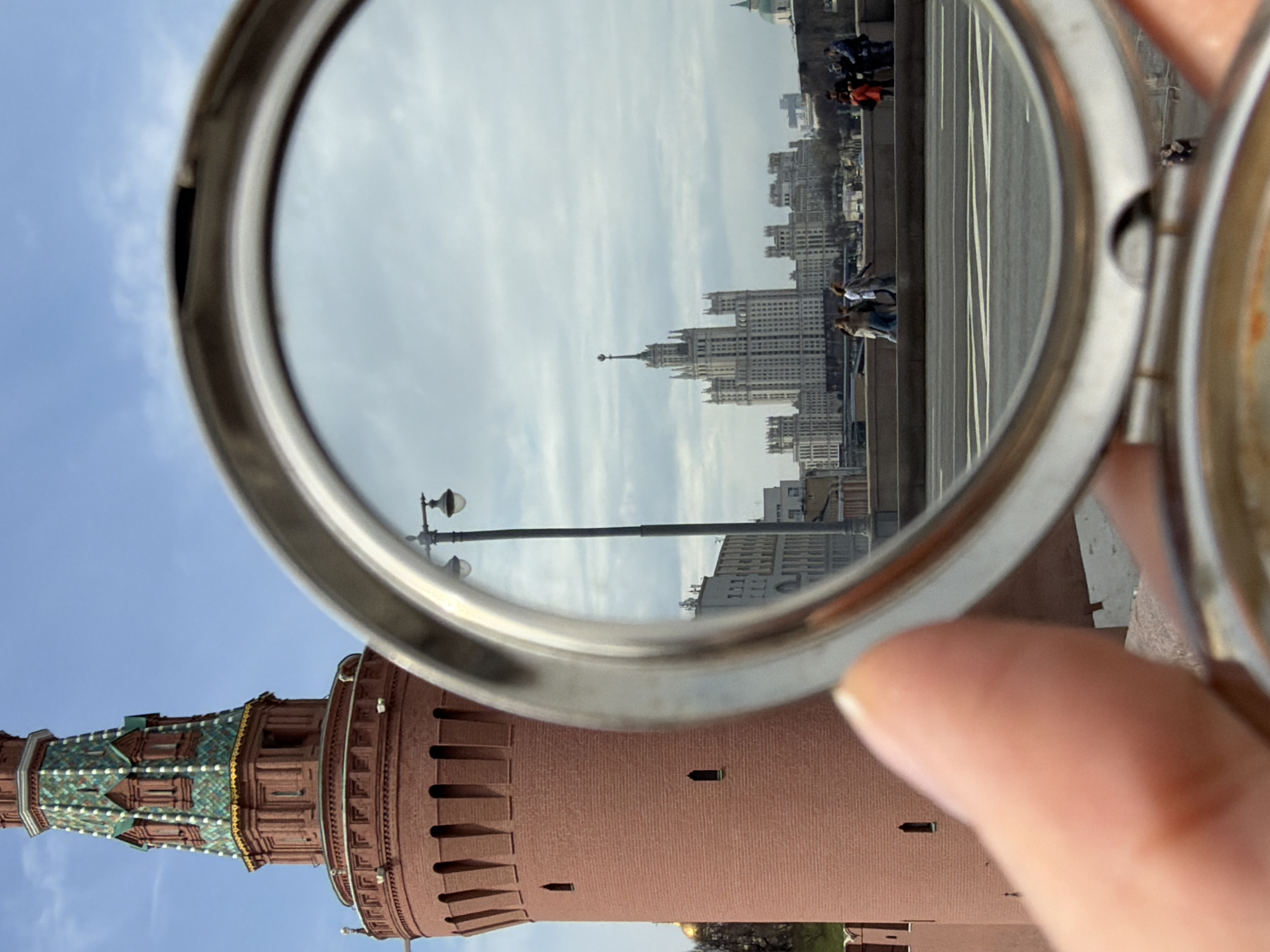 A partial view of the Moscow Kremlin is seen in the foreground, with a focus on one of its towers, distinguished by its brick exterior and characteristic green-tiled roof. In the background, reflected in a metallic, round-shaped mirror held by a person, is a cityscape. The reflection shows a line of buildings, with one prominent, tall skyscraper visible among the others. A bridge is also reflected, with figures of people walking along it. The sky is partly cloudy. The scene is an outdoor setting.