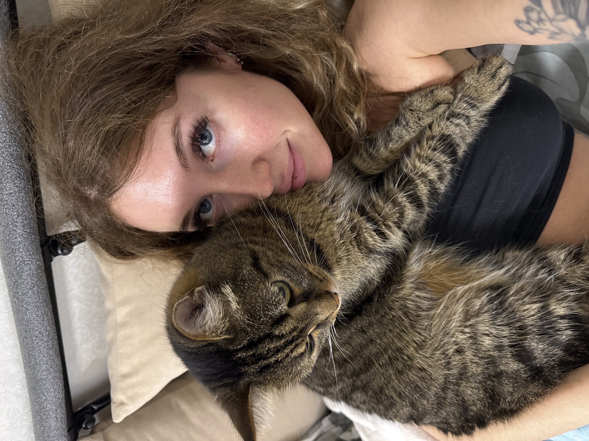 The image shows a close-up, overhead shot of a woman and her cat. The woman, with long, wavy blonde hair and blue eyes, is lying down, her face angled towards the camera with a slight smile. She is wearing a black top. A tabby cat is nestled close to her, its head resting on her chest. The cat's fur is a mix of brown, black, and grey tabby patterns, and it has green eyes looking slightly off-camera. The scene appears to be indoors, possibly a bedroom, with soft lighting. The woman has a tattoo on her upper arm. The overall mood of the image is one of comfort and affection between the woman and her pet.