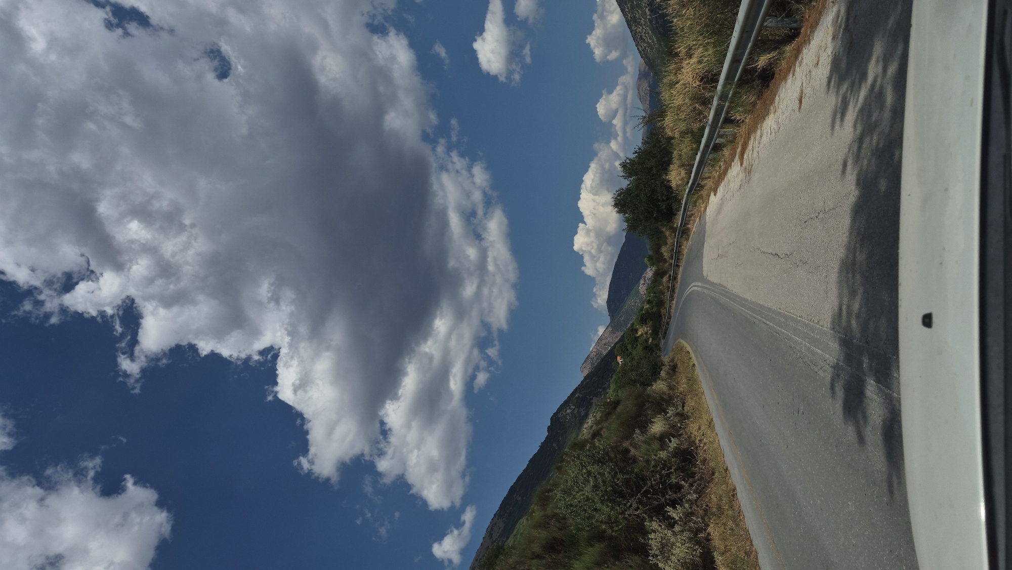A low-angle, vertical, full shot captures a view from inside a vehicle driving on a winding asphalt road through a mountainous, rural landscape under a partly cloudy blue sky. The road, which appears to be a two-lane highway, curves to the left and disappears into the distance. The right side of the road is lined with a metal guardrail, beyond which are dry, yellowish grasses and bushes. To the left of the road, a steep, green, vegetation-covered slope rises. In the distance, rolling hills and mountains are visible, some with sparse vegetation. The sky is a vibrant blue, dotted with large, fluffy white and gray clouds. The foreground shows the edge of a white vehicle's dashboard and windshield. The scene is illuminated by bright daylight, casting tree shadows on the road. There are no people visible in the image. The location is not specified, but the environment suggests a warm, possibly Mediterranean climate.