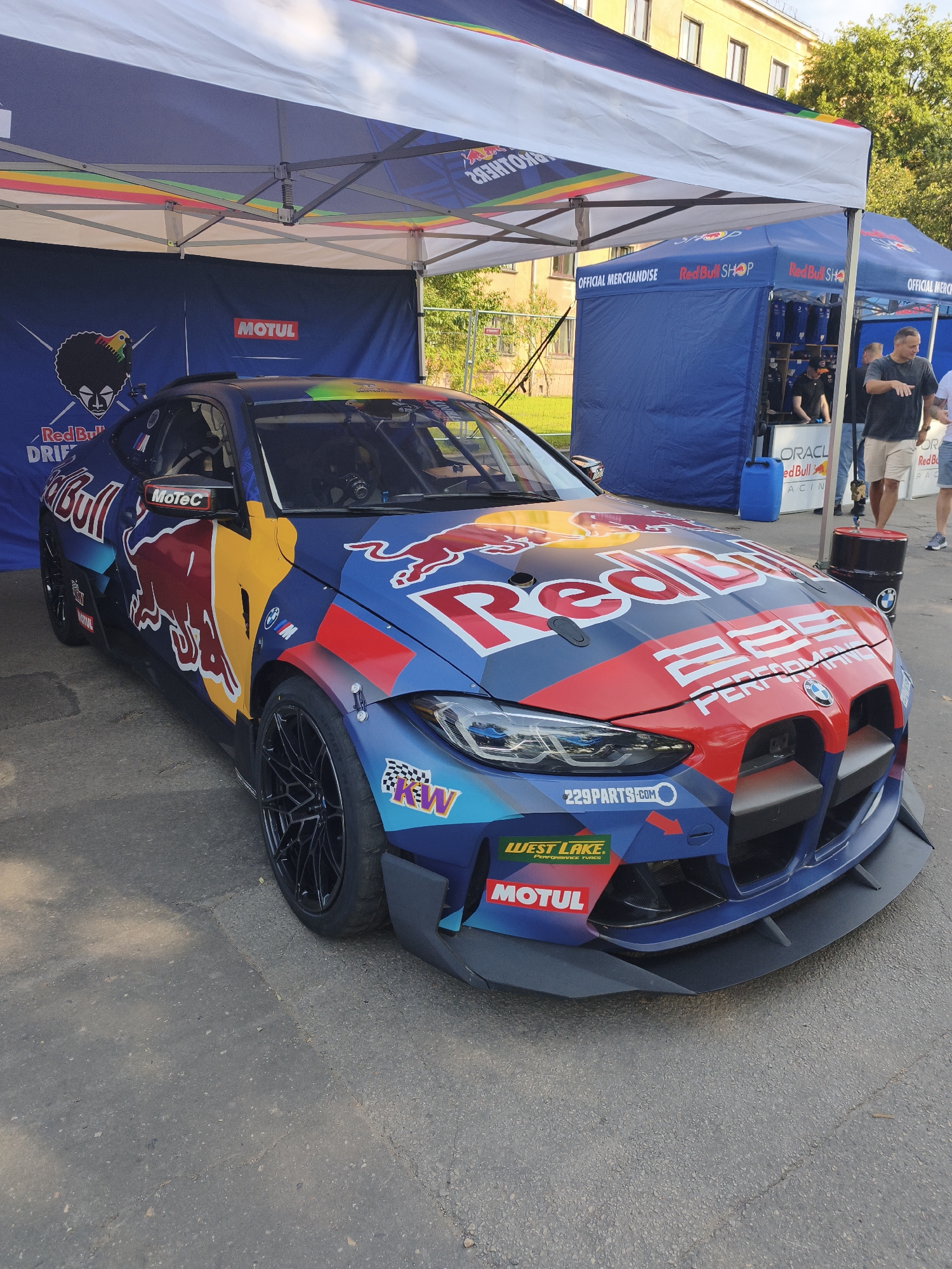 The image depicts a vibrant, eye-catching racing car, likely a BMW M4, prominently displayed outdoors under a white tent with colorful striped accents. The car is adorned with the iconic Red Bull logo and other racing sponsorships, including Motul and West Lake Performance Tyres. The racing car is parked on an asphalt surface, which appears to be a street or public area. In the background, there are blue tents that seem to be part of a Red Bull merchandise or event setup. One of these tents has "OFFICIAL MERCHANDISE" and "Red Bull SHOP" signage. Beyond the tents, a building with windows and trees are visible, suggesting an urban setting. Several people are present, some standing near the merchandise tents, and others are blurred in the background. The lighting indicates it is daytime, with clear sky. The overall atmosphere suggests an event or promotional activity related to motorsports. The presence of Latvian flags on the car further confirms the location in Riga, Latvia.