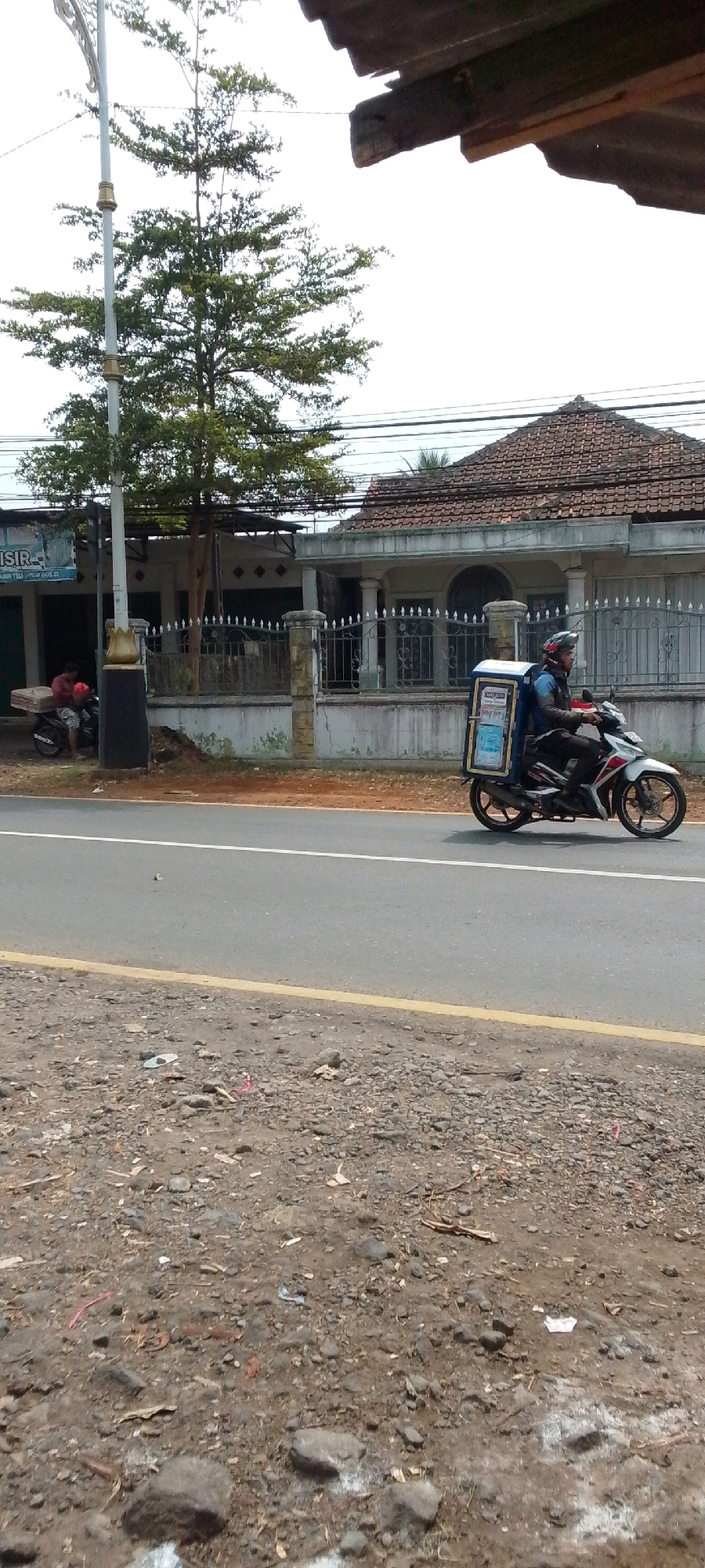 In Cilaku, Indonesia, on a moderately overcast day, a person is riding a white and red motorcycle. The rider, wearing a blue jacket, black pants, and a helmet, has a blue insulated delivery box mounted on the back of the motorcycle. The motorcycle is traveling on an asphalt road, which has a single yellow line marking the shoulder. On the left side of the road, a paved area with dirt and gravel is visible. Behind the road and the paved area, a building with a tiled roof and a fence is present. A tree with green foliage stands near a lamppost in front of the building. To the left of the tree, another motorcycle is parked, with a person in a red shirt visible beside it. The scene appears to be along a roadside in a semi-urban or residential area.