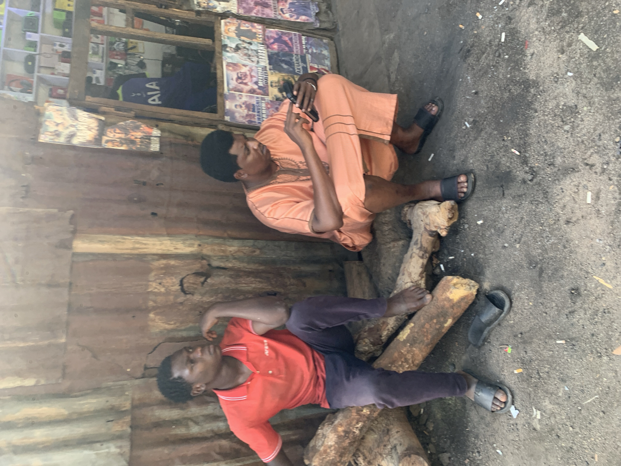 Two men are sitting outside a building in Jos, Nigeria. One man is sitting upright, wearing an orange outfit and looking at a phone. The other man is lying down on a pile of logs, wearing a red shirt and dark pants. Both men are wearing sandals. The ground around them is littered with cigarette butts and trash. The building behind them has a corrugated metal wall and a window displaying various items, including movie posters and a Tottenham Hotspur jersey.