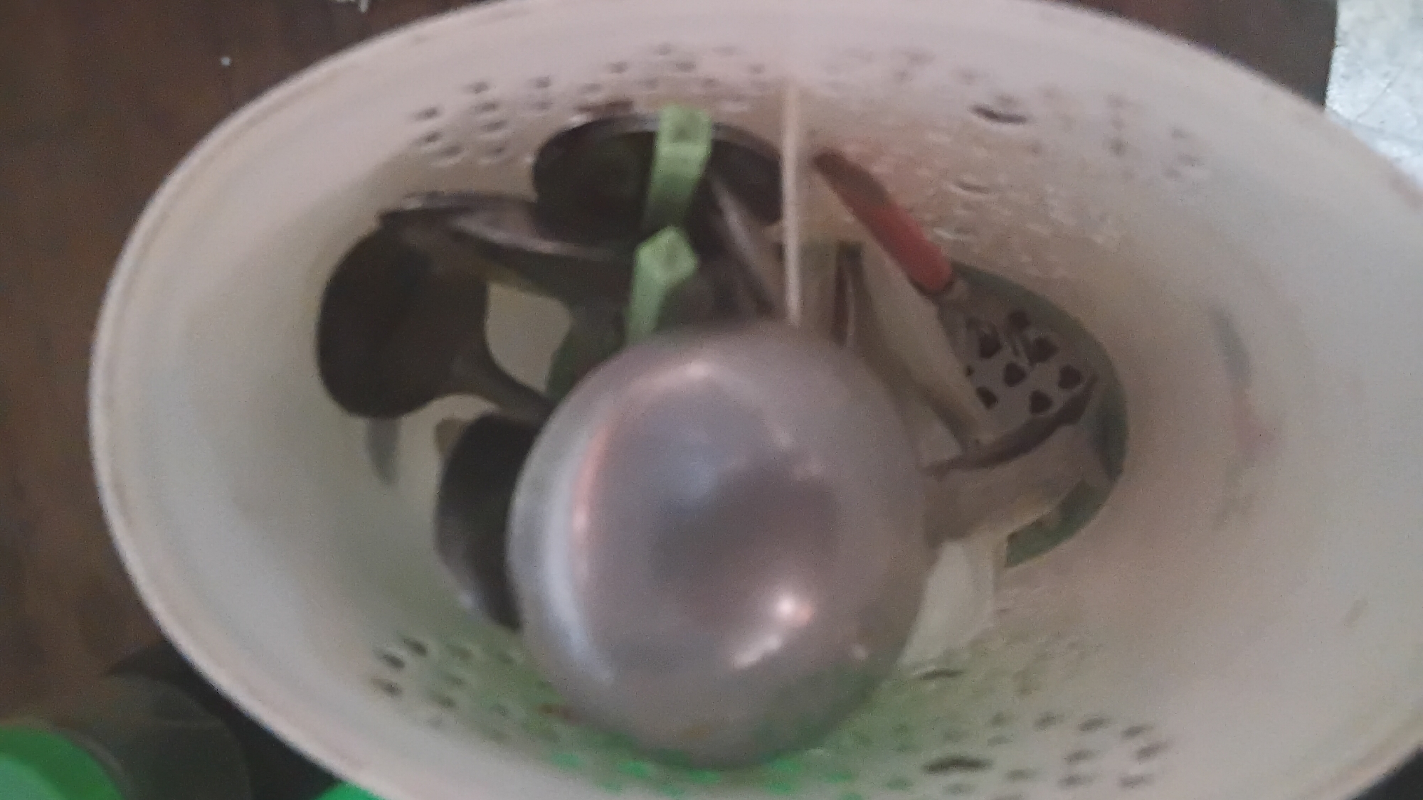 A high-angle, close-up shot shows a collection of kitchen utensils, including ladles and spoons, stored in a white plastic basket. The basket is perforated with circular holes, allowing visibility of the utensils within. A large, silver ladle with a rounded bowl dominates the foreground, positioned to the left of the frame. To the right, several darker-colored spoons with curved handles are visible, along with two green plastic tongs. A small red utensil handle is also discernible at the bottom right. The background consists of a blurry brown surface, likely a tabletop. The scene is set in a kitchen environment, possibly in Prabumulih, Indonesia. The lighting is dim and diffused, resulting in a slightly soft focus across the image. No individuals are present in the frame.