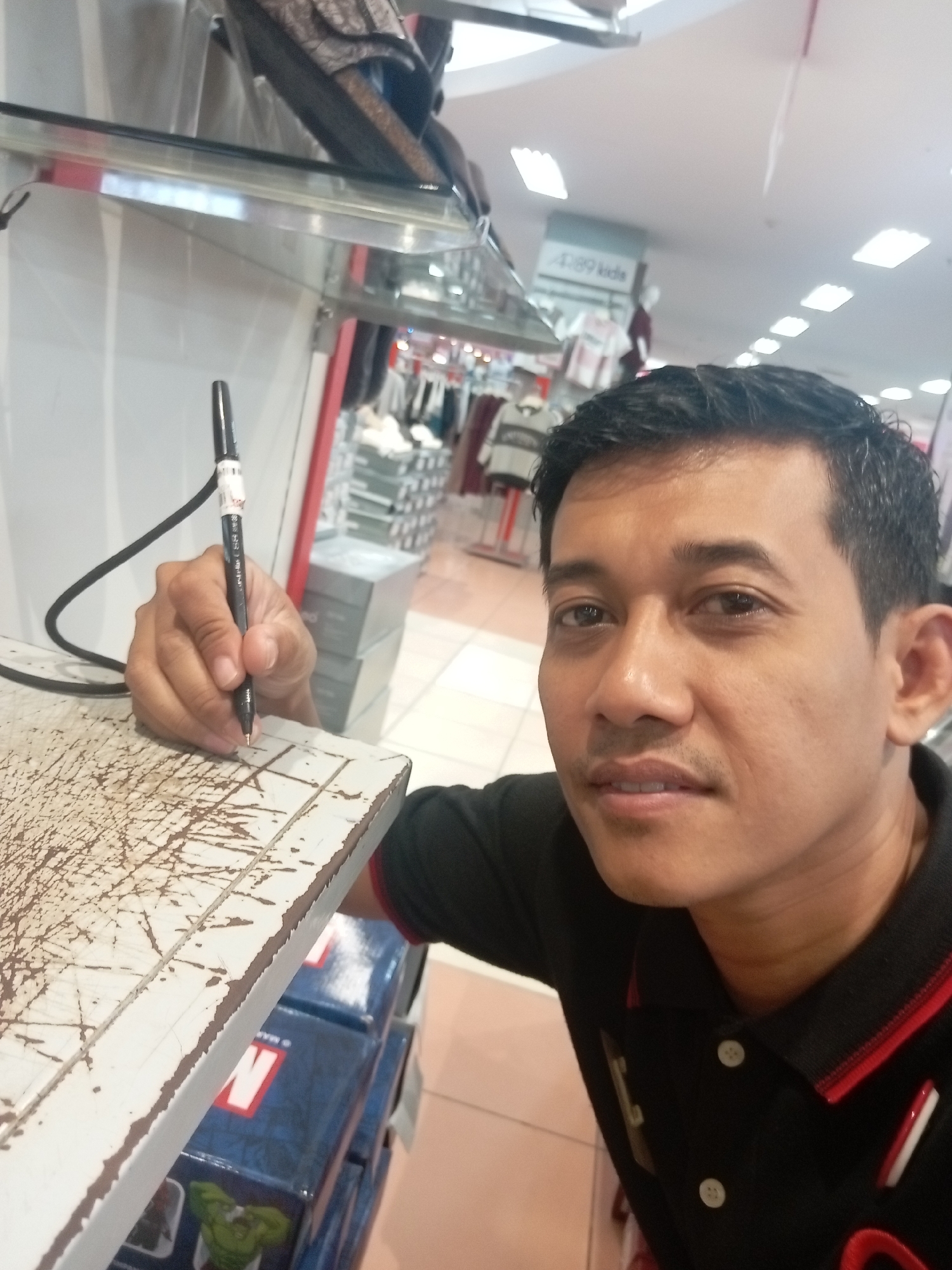 A man with short, dark hair and a mustache is taking a selfie in what appears to be a retail store. He is wearing a black polo shirt with red trim and white buttons. He is holding a black pen in his right hand and appears to be writing on a white surface with brown markings. In the background, shelves stocked with merchandise and a sign with "d89 kids" are visible. The lighting suggests an indoor environment, possibly a mall or department store in Batam, Indonesia.