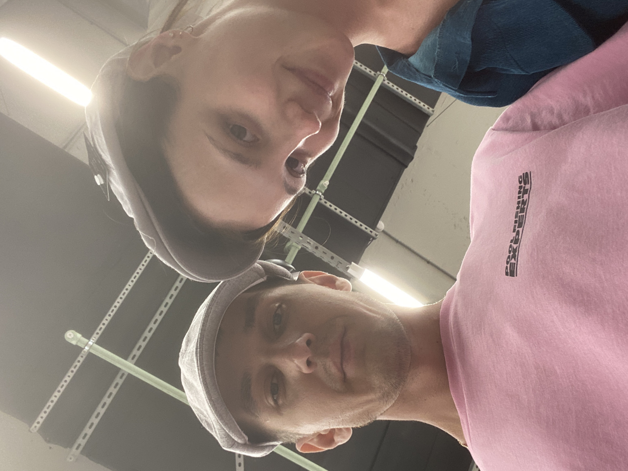 This is a low-angle, close-up selfie of two individuals, a man and a woman, wearing flat caps. The man is on the left, wearing a pink t-shirt with black text that appears to read "POOL FISHING EXPERTS". The woman is on the right, wearing a blue top. They are looking downwards towards the camera. The background consists of a ceiling with industrial lighting and a metal structure. The scene appears to be indoors, possibly in a retail or storage space. The presence of industrial lighting and shelving suggests a commercial or service-oriented environment. No specific identifying features of Novokuybyshevsk, Russia are visible.