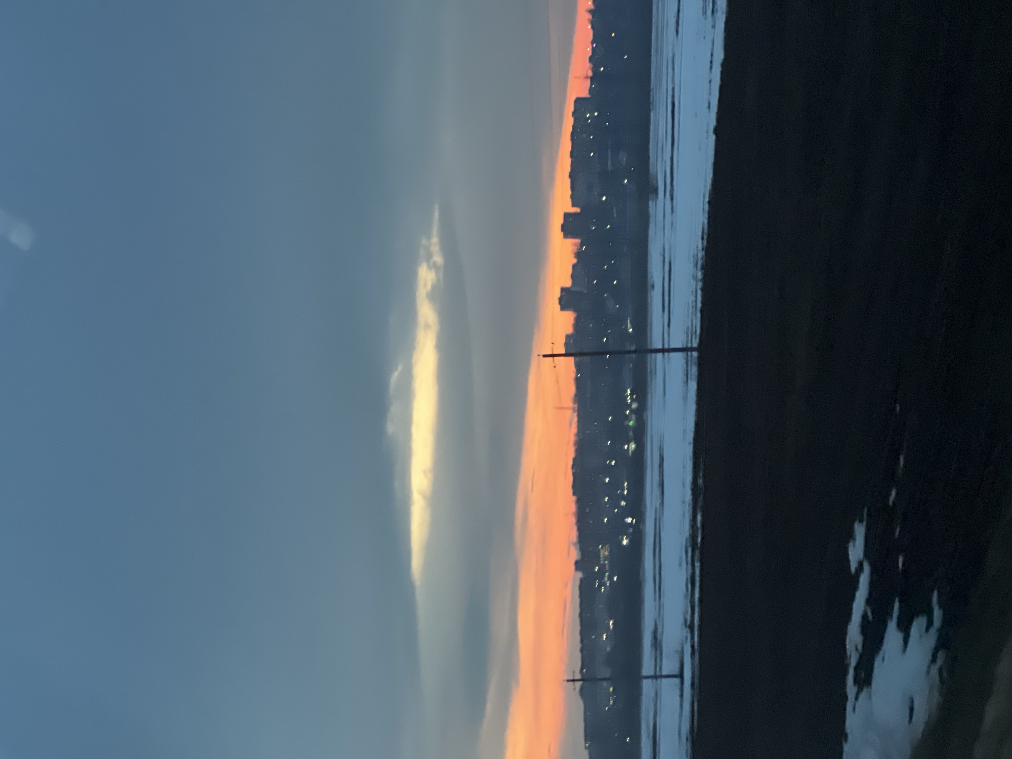 The image depicts a view in Votkinsk, Russia, likely at sunset or twilight. To the left, the sky transitions from a blue-gray to orange hues towards the horizon. A thin, bright cloud is visible in the left center. In the middle ground, a silhouette of the city skyline is visible along with lights of the buildings and street lights. Power lines or poles are present, silhouetted against the colorful sky. To the right, a dark land mass or shoreline borders a body of water that reflects the city lights. The lighting suggests evening or early morning. The image is oriented sideways.