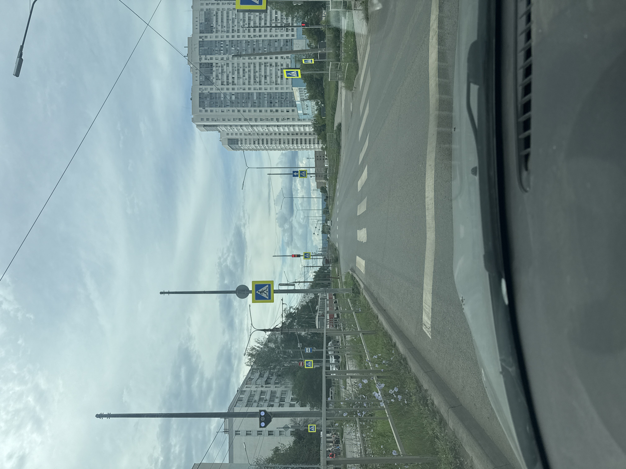 The image captures a view from inside a vehicle looking forward along a city street in Samara, Russia. The overcast sky suggests a cloudy day. The street is paved and features multiple pedestrian crossings marked by white stripes. On the left side of the road, a metal fence separates the sidewalk from a grassy area with some small blue wildflowers. Beyond the fence, a multi-story apartment building with numerous windows and balconies lines the street. Lampposts and traffic signals are visible, along with several pedestrian crossing signs. One traffic light shows a blue signal. Further down the street, a red tram can be seen. On the right side of the road, a very tall, modern apartment building dominates the skyline, also with many windows. Power lines are strung across the sky. The vehicle's dashboard and windshield frame the scene.