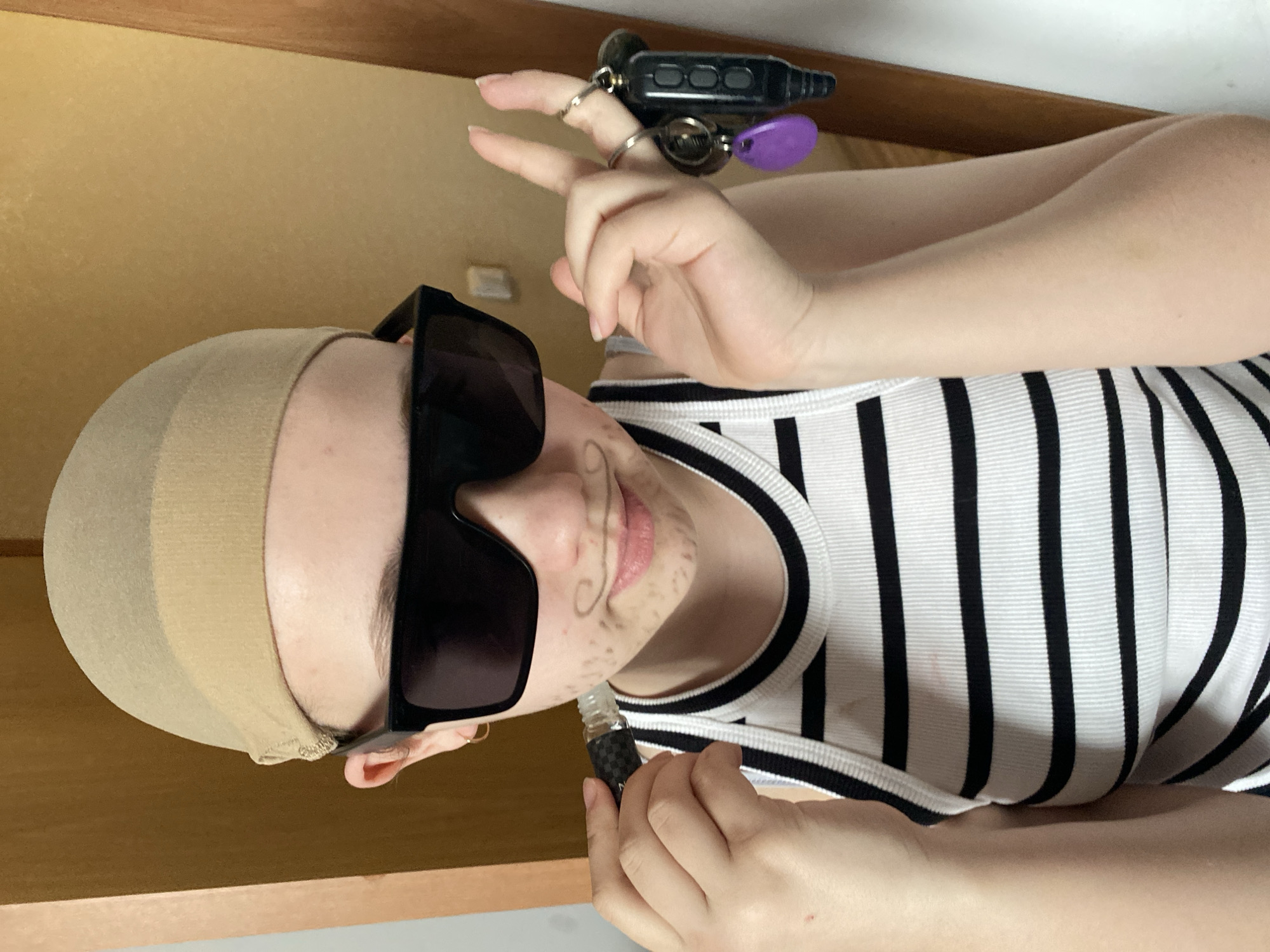 The image shows a person wearing a tan head covering, oversized black sunglasses, and a white tank top with black vertical stripes. The person has drawn a mustache and stubble on their face. They are holding a small vial in their right hand and a set of keys in their left hand, making a peace sign. The background appears to be a room with wooden cabinets.
