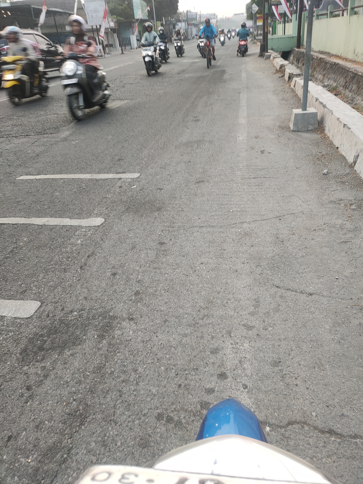 The image depicts a street scene in Kanigoro, Indonesia, with multiple motorcycles and a bicycle navigating the asphalt road. Several individuals are riding motorcycles, some wearing helmets and casual attire. A bicycle is also visible with a rider. The road is marked with white lines, and a concrete drainage ditch runs along the right side of the street, bordered by a low concrete wall. Buildings and flags are visible in the background. The perspective suggests the image was taken from the viewpoint of someone riding a motorcycle. The overall environment appears to be an urban or semi-urban setting with moderate traffic.