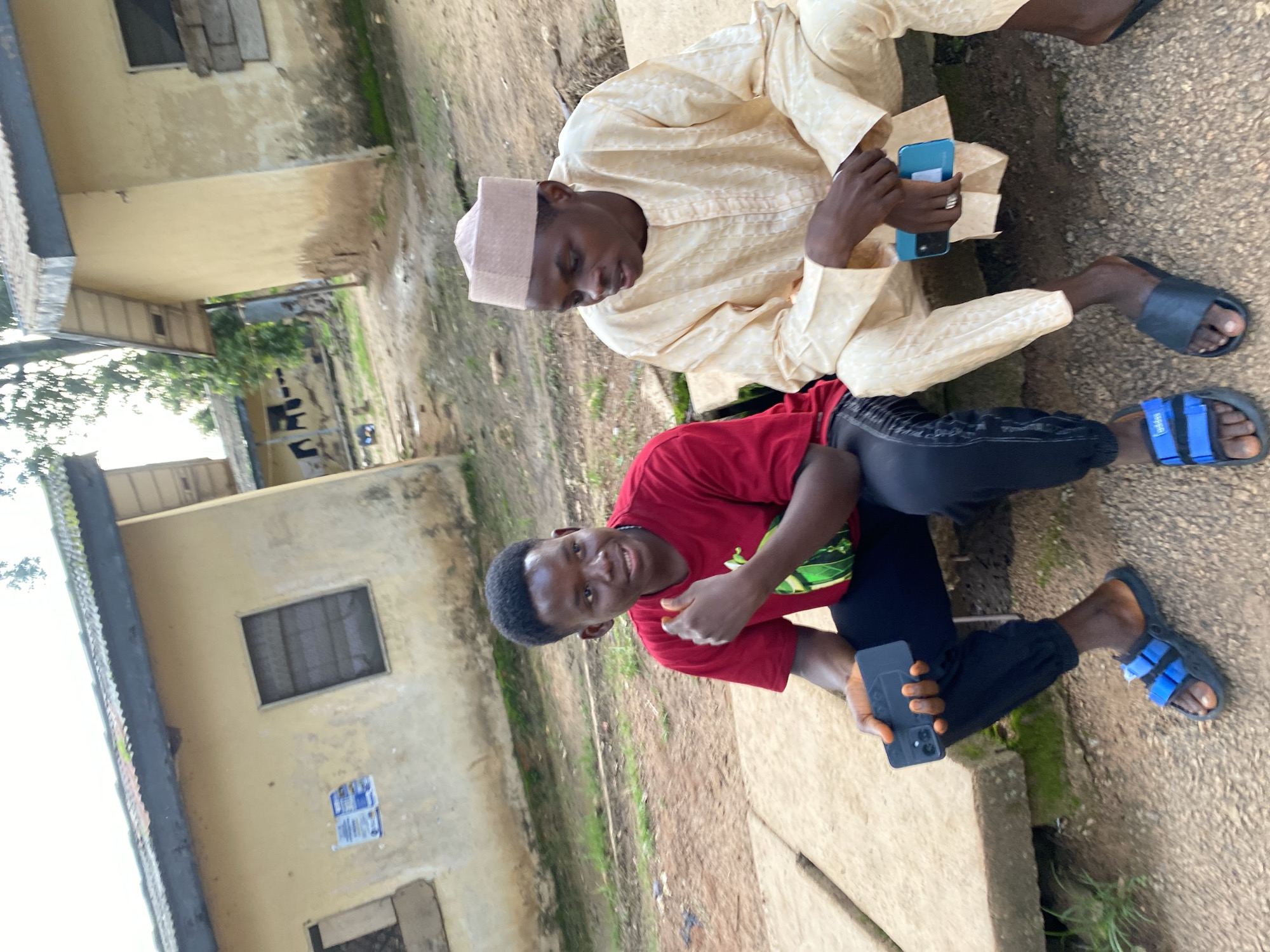 Two young men are sitting on a concrete ledge outdoors. The man on the left is wearing a red t-shirt and black pants, and is holding a black smartphone. He is wearing blue and black sandals. The man on the right is wearing a light yellow traditional outfit and a light brown cap. He is holding a light blue smartphone. Both men appear to be looking towards the viewer or slightly to the right. In the background, there are two weathered buildings with peeling paint, corrugated metal roofs, and barred windows. The ground is covered in gravel and dirt, with some patchy grass. The overall scene suggests a casual outdoor setting, possibly in a rural or semi-urban area in Rafin Par, Nigeria, given the architectural style of the buildings and the traditional clothing worn by one of the individuals.