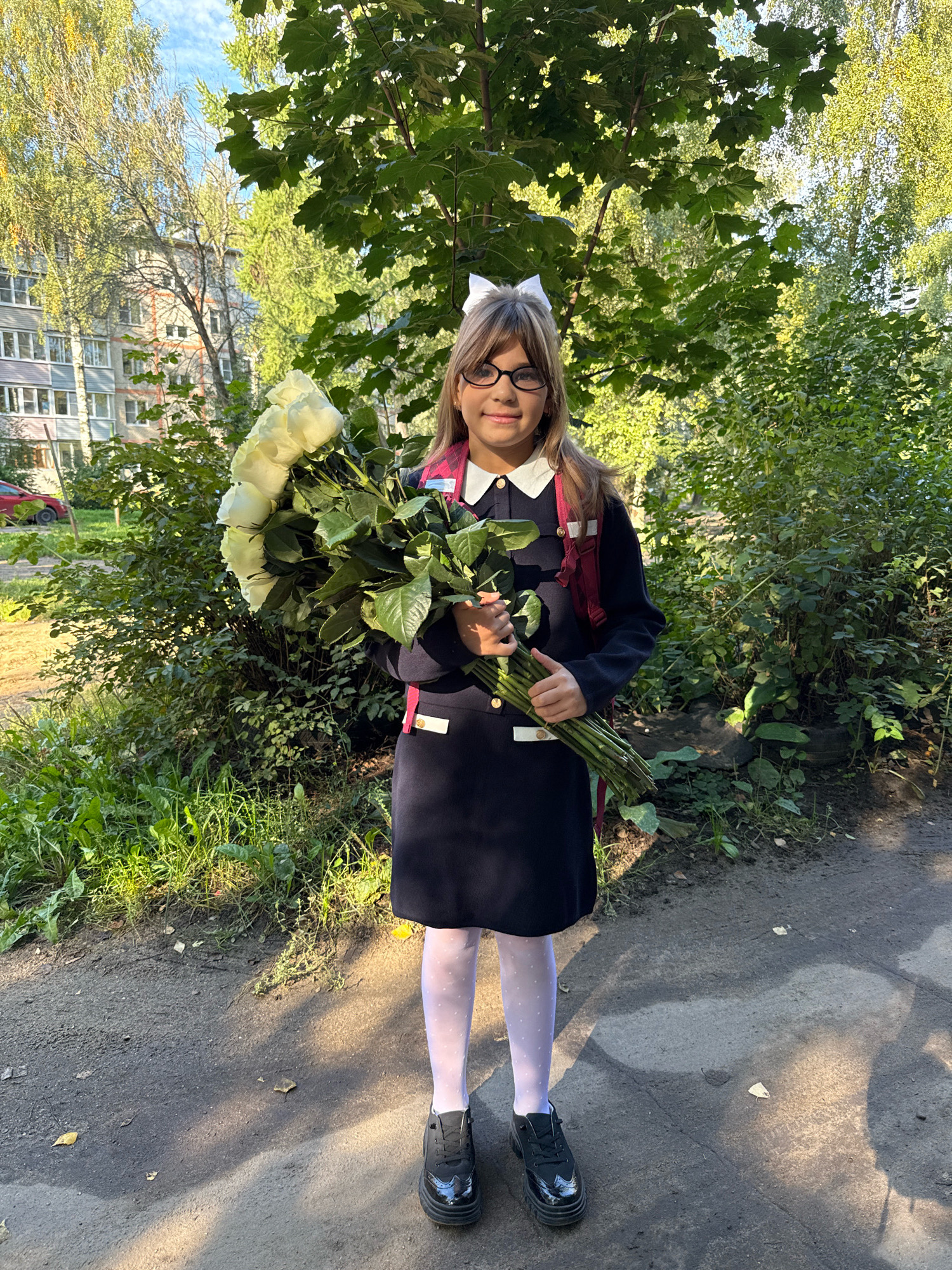 A young girl with blonde hair, wearing a navy blue school uniform with a white collar and a large white bow, stands outdoors holding a bouquet of white roses. She wears black-rimmed glasses, white polka-dot tights, and black patent leather shoes. A maroon backpack is visible on her back. She is positioned in front of a lush green bush with a building and trees in the background. The ground in front of her is a dirt path with scattered leaves. The scene appears to be in a residential area, likely in or near the city of Rusanovo, Russia, given the architectural style of the building and the surrounding vegetation. The lighting suggests it is daytime, possibly late afternoon, with sunlight casting shadows on the ground.