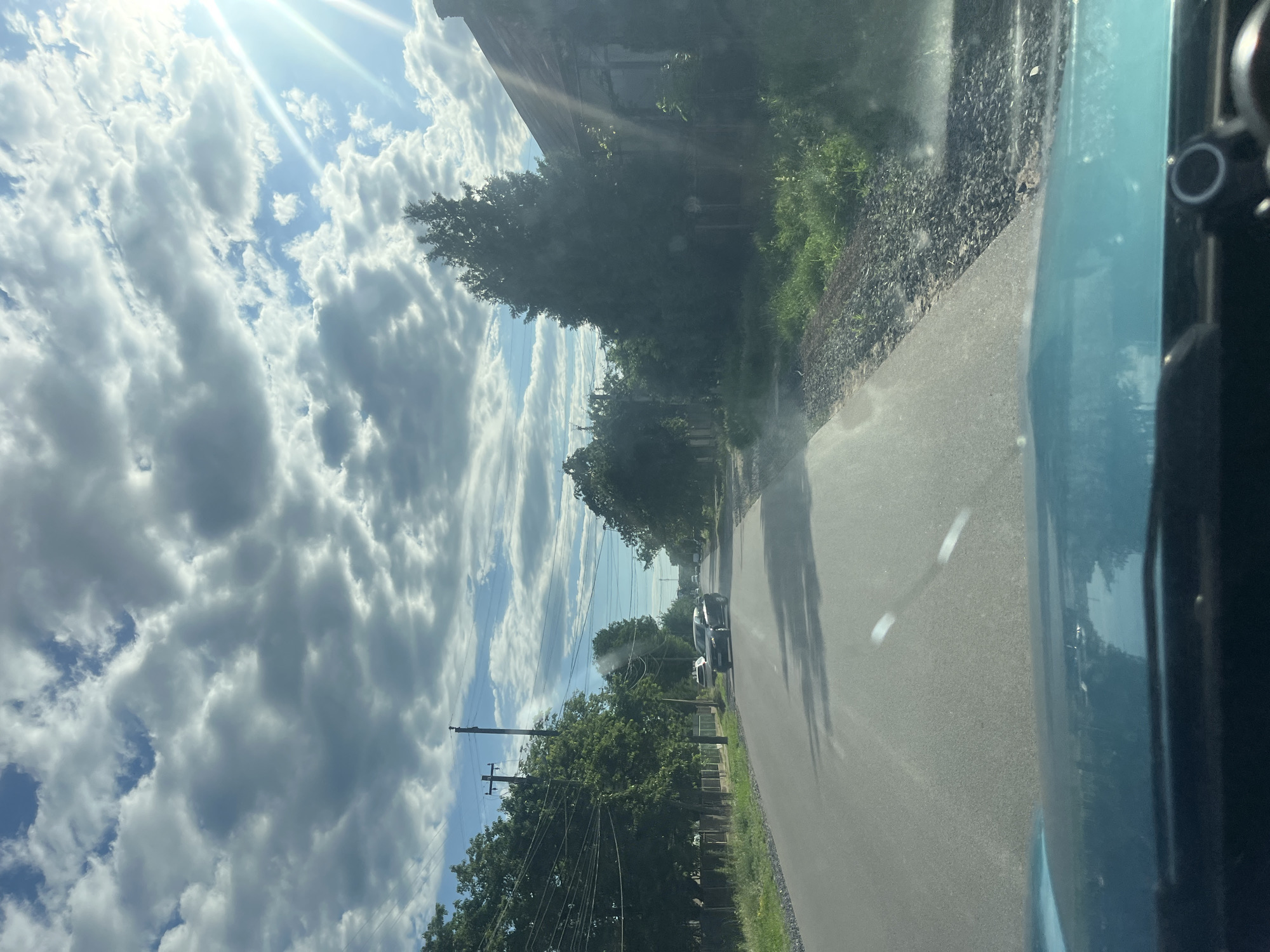 This image depicts a view from inside a vehicle traveling on a paved road in Storozhnytsia, Ukraine. The road is lined with green trees and vegetation on both sides. On the left, there is a wooden fence and utility poles with numerous overhead wires. On the right, an old, weathered building with a dark roof is visible, partially obscured by a tall evergreen tree. Several cars are present on the road; one dark sedan is in motion, and another white vehicle is parked further ahead. The sky is partly cloudy, with dramatic, textured clouds and bright sunbeams piercing through from the upper right corner. The scene appears to be in a rural or suburban area, characterized by a mix of natural elements and infrastructure.