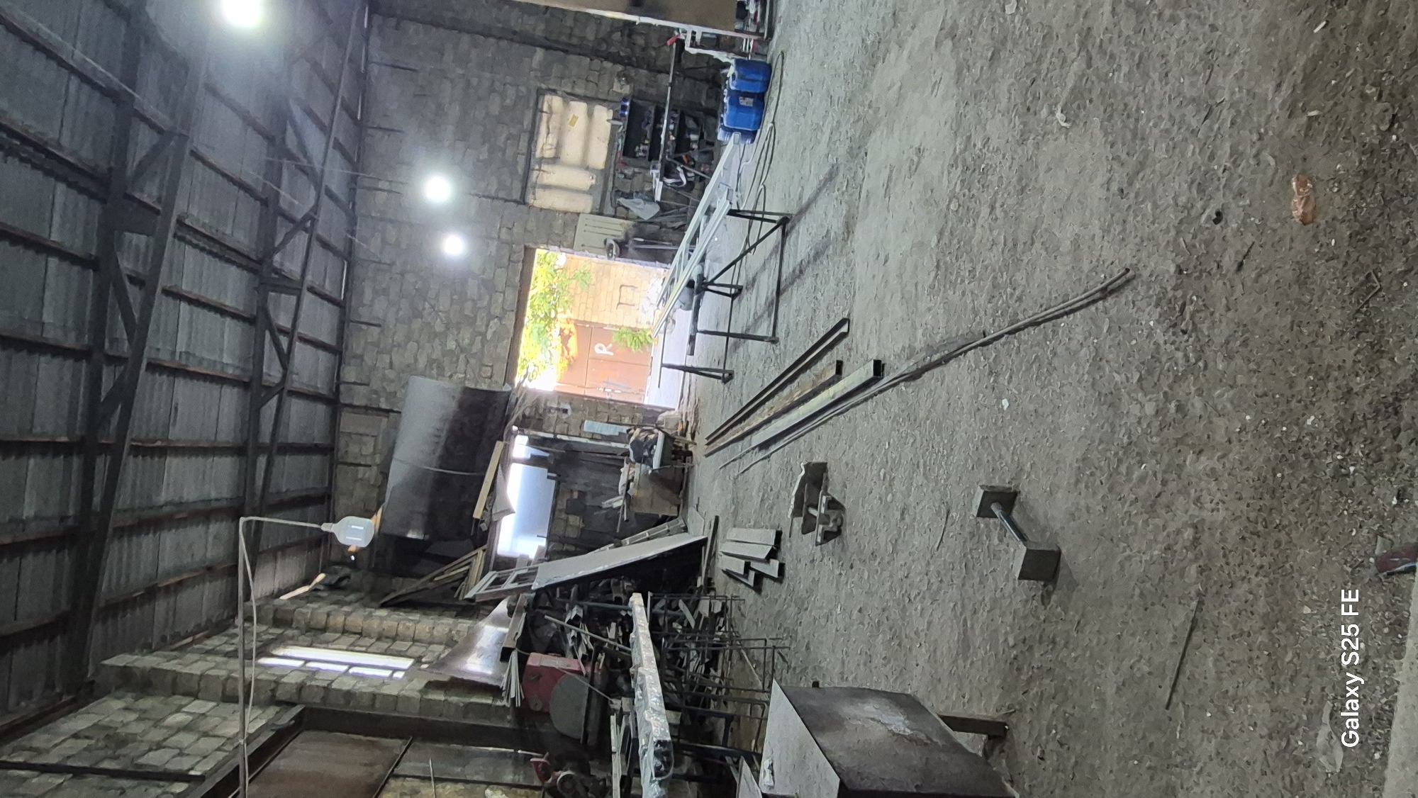 The image depicts the interior of a workshop or storage facility in Bilajari, Azerbaijan. The space has a rough, industrial feel, with exposed stone walls and a corrugated metal roof supported by wooden beams. The floor is made of dirt and is cluttered with various objects.
In the foreground, a metallic dumbbell-like object rests on the ground. Further back, several long metal beams are laid out on the floor, along with some smaller metal pieces. To the left, a workbench is visible, laden with tools and materials, and a large metal container is positioned in the lower left corner.
The back of the workshop opens up to an outdoor area, possibly a yard or street, with natural light streaming in. Two large blue barrels are placed near the entrance. The overall impression is of a working environment, possibly for metal fabrication or storage. The presence of the "Galaxy S25 FE" watermark at the bottom left suggests the image was captured using a Samsung smartphone.