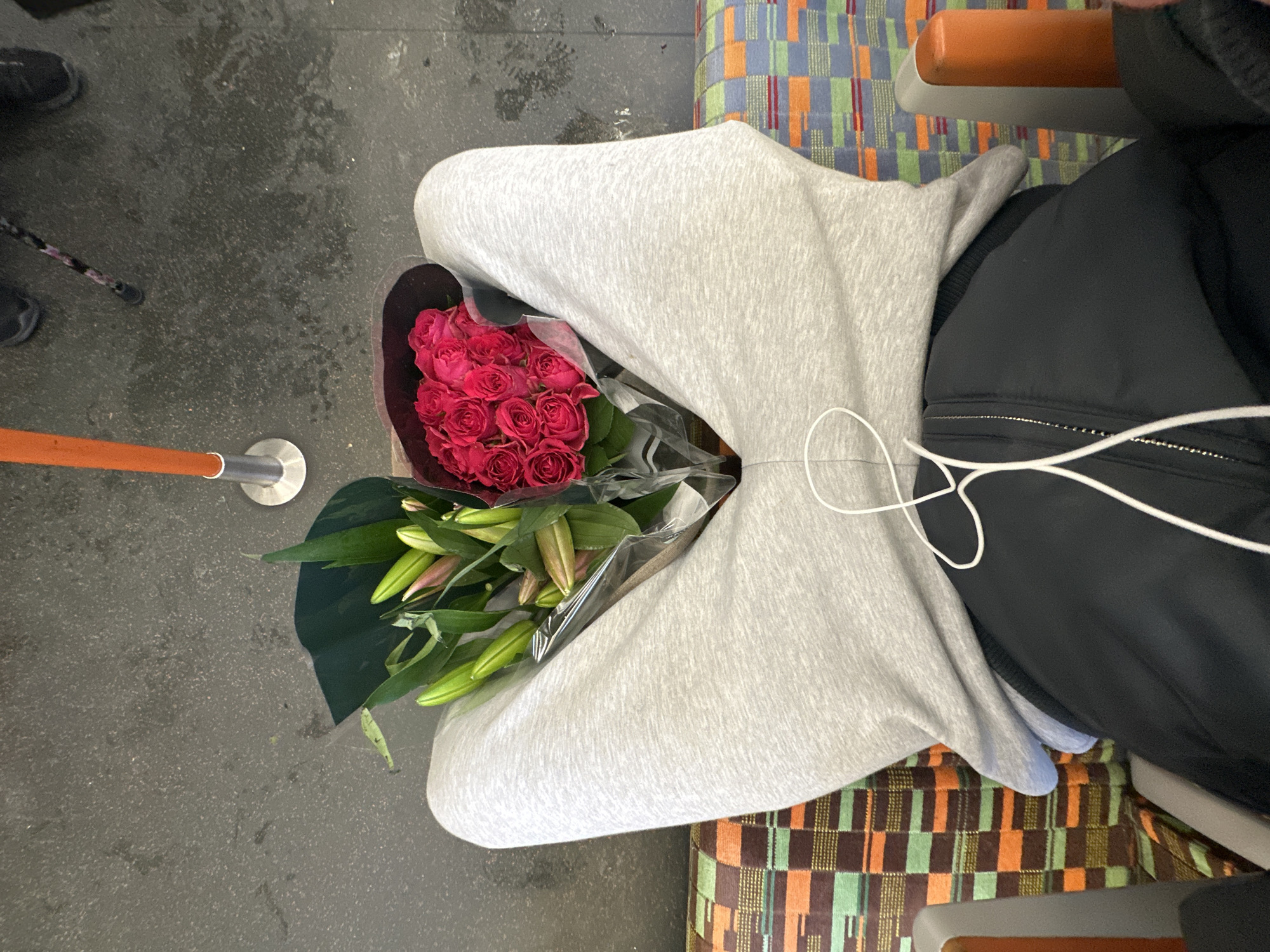 A top-down, eye-level shot shows a person seated on a train, holding a bouquet of flowers. The person is wearing a light grey hoodie, with the hood spread open to cradle a bouquet of roses and lilies in a maroon wrapping. The person is also wearing a black jacket with white drawstrings. The seat has a colourful geometric pattern. The train floor is visible in the periphery, and an orange pole. Given the seat pattern and the presence of the orange pole, this is likely a train on the London Underground, United Kingdom.