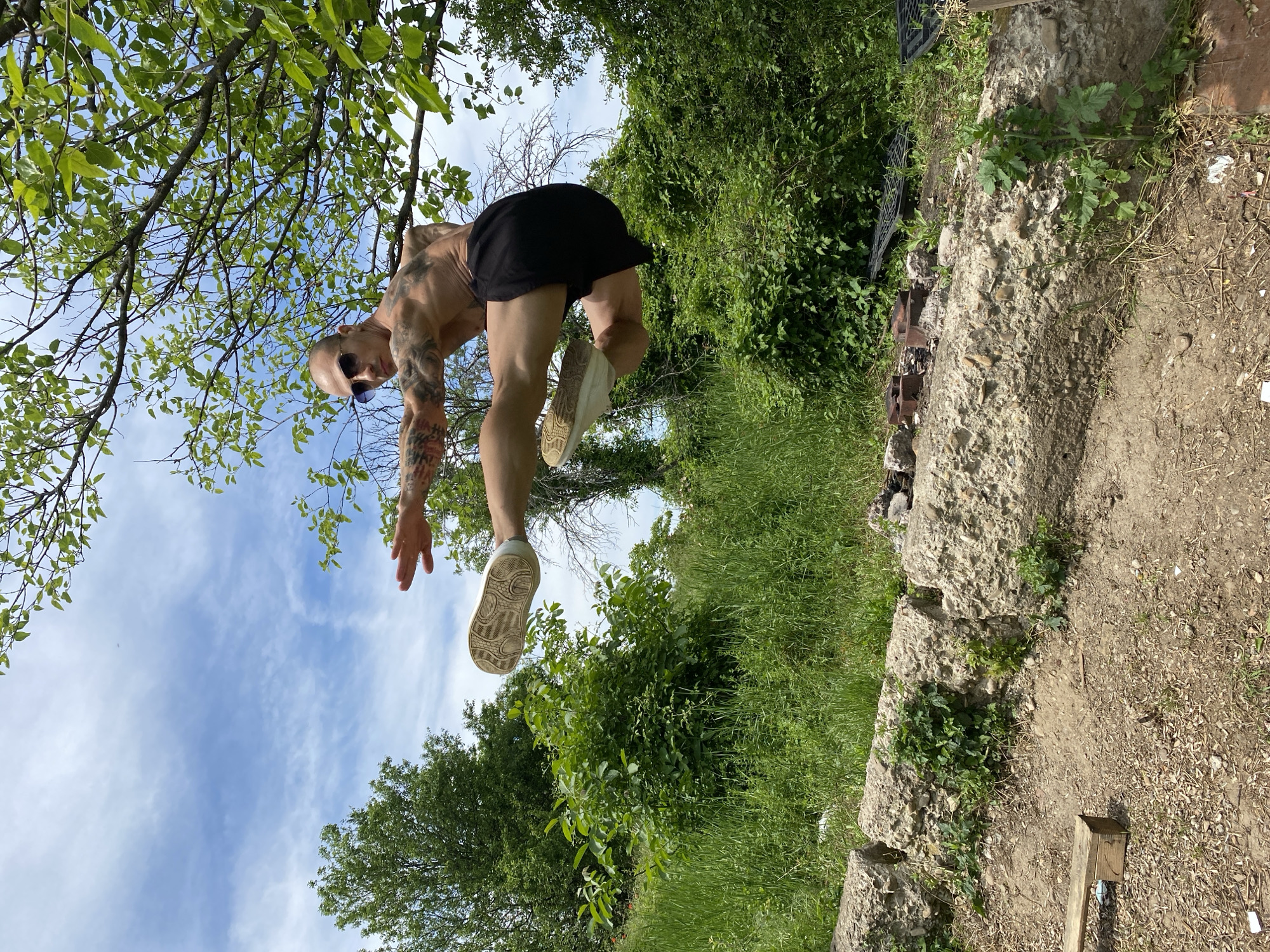 A person is jumping outdoors. The person, likely a male with tattoos on the arms, is wearing shorts and white shoes. The background includes trees, sky, and a stone structure. The location is Pleven, Bulgaria.