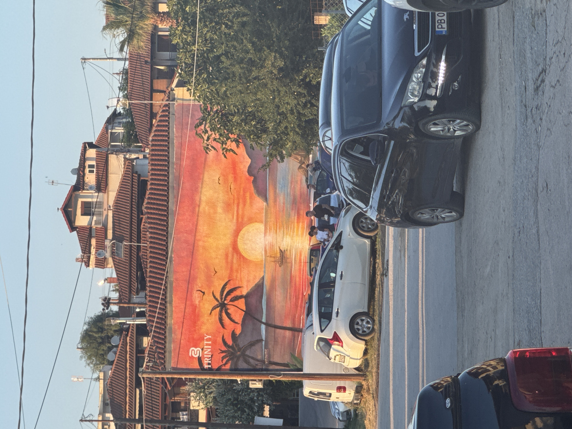 The image depicts a street scene in Ammouliani, Greece. Dominating the background is a building adorned with a large mural of a sunset over the sea, featuring palm trees, islands, and a sailboat. The mural has the word "TRINITY" prominently displayed with a logo above it. In the foreground, several cars are parked along the roadside. A white hatchback is parked to the left, next to a white van. A black station wagon is parked in the center, facing right, with its headlights illuminated. Another black vehicle is partially visible in the lower left corner. A few individuals are standing near the white hatchback, some appearing to be interacting. The environment suggests an outdoor setting, likely during the daytime with clear skies, as indicated by the bright sunlight and shadows. The presence of parked cars, buildings, and overhead wires points to a developed urban or semi-urban area.