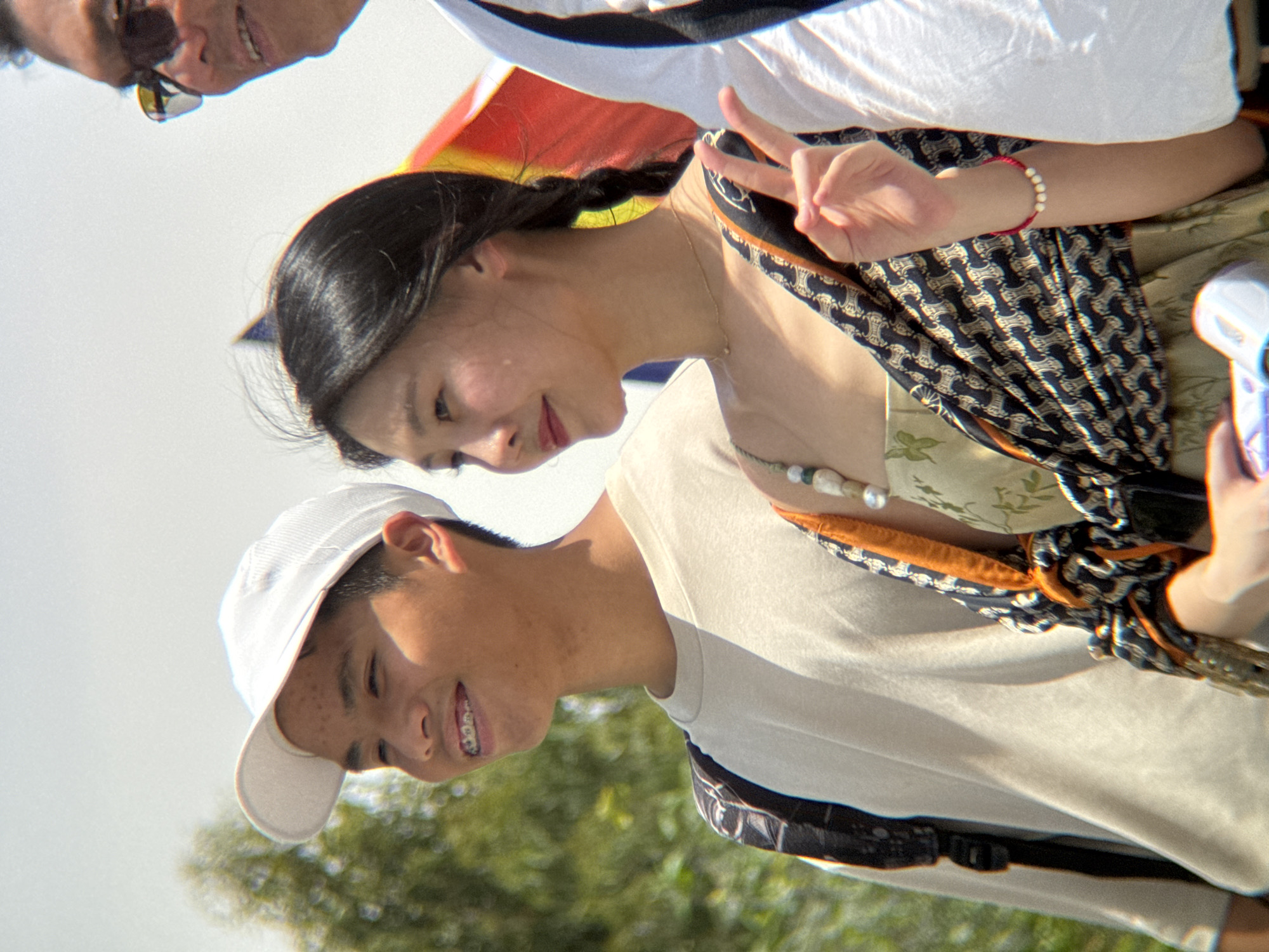 A young man wearing a white baseball cap and a light-colored t-shirt stands to the left. He has braces and is smiling. He is wearing a backpack. To his right, a young woman with dark hair styled in a braid wears a patterned scarf and a light green dress. Her hand is raised, making a peace sign. She is wearing a beaded necklace and a bracelet. Behind them, a man in sunglasses is partially visible. A rainbow-colored flag is also visible in the background. The scene appears to be outdoors, possibly in a city, with trees and a bright sky suggesting a sunny day. The overall atmosphere seems festive. There are no definitive visual cues in the image to confirm the location as Nha Trang, Vietnam.