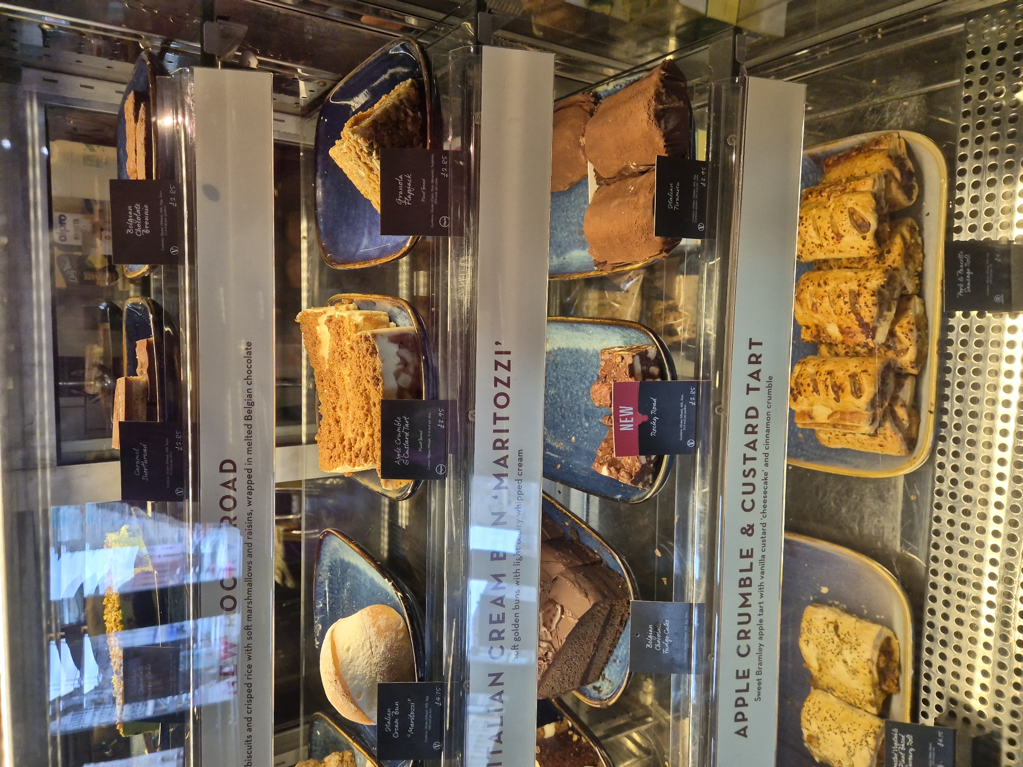 The media file captures a well-lit refrigerated display case filled with an assortment of cakes, tarts, and pastries, likely in a café or bakery. The scene is shot from a slightly elevated, eye-level perspective, looking down into the case, which is divided by vertical clear plastic dividers holding product information. Many items are presented on blue or white rectangular plates and trays.
Visible food items include individual slices of cake, square bars, buns, and sausage rolls. Among the identifiable products are:
- Caramel Shortbread, priced at £2.85.
- Belgian Chocolate Brownie, also £2.85.
- Granola Flapjack, marked as "Plant Based" and costing £2.85.
- Italian Tiramisu, priced at £3.95.
- Belgian Chocolate Fudge Cake.
- Rocky Road, labeled "NEW" and costing £2.85, described as "biscuits and crisped rice with soft marshmallows and raisins, wrapped in melted Belgian chocolate."
- Italian Cream Bun "Maritozzi", priced at £4.15, described as "Soft golden buns with light airy whipped cream."
- Apple Crumble & Custard Tart, with one version marked "Plant based" at £3.95, and a general description stating "Sweet Bramley apple tart with vanilla custard 'cheesecake' and cinnamon crumble."
- Pork & Pancetta Sausage Roll, with an adjacent label mentioning "Plant Based" and £4.15, suggesting a plant-based alternative or a price for another item nearby.
The prices are in British Pounds (£), indicating a UK location, consistent with the provided context of West Suffolk. Some labels also include dietary information, such as "Plant Based" or "V" (vegetarian, assumed for the "Belgian Chocolate Brownie" and "Caramel Shortbread" based on similar labels). The brand "alpro" is partially visible in the background. The display appears clean and well-organized, with items presented neatly. No people are visible in the frame.